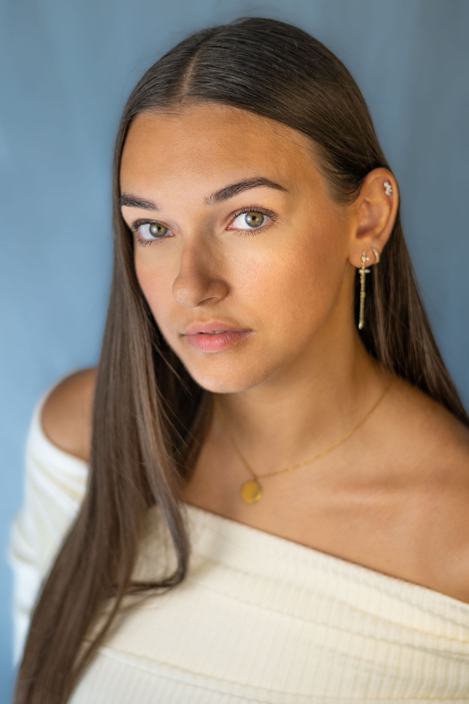 Portrait series in off-shoulder white top with gold accessories against blue backdrop.