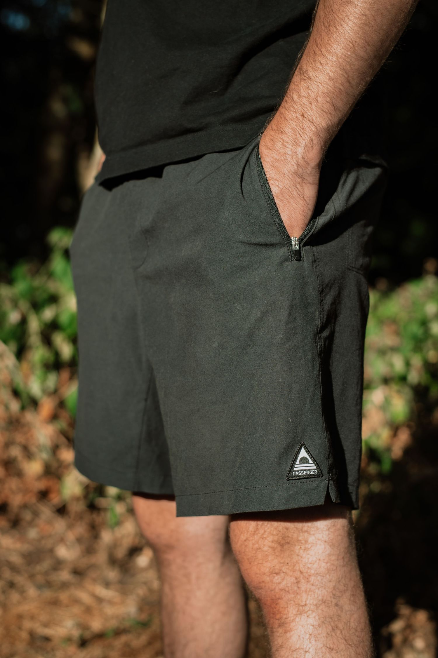 Man wearing dark olive green athletic shorts with zippered pocket and triangle logo patch outdoors.