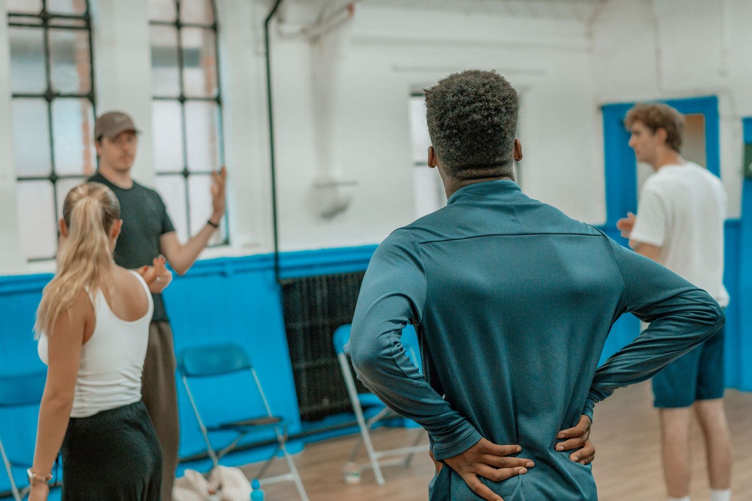 Group of performers rehearse choreography in a spacious dance studio with natural lighting.