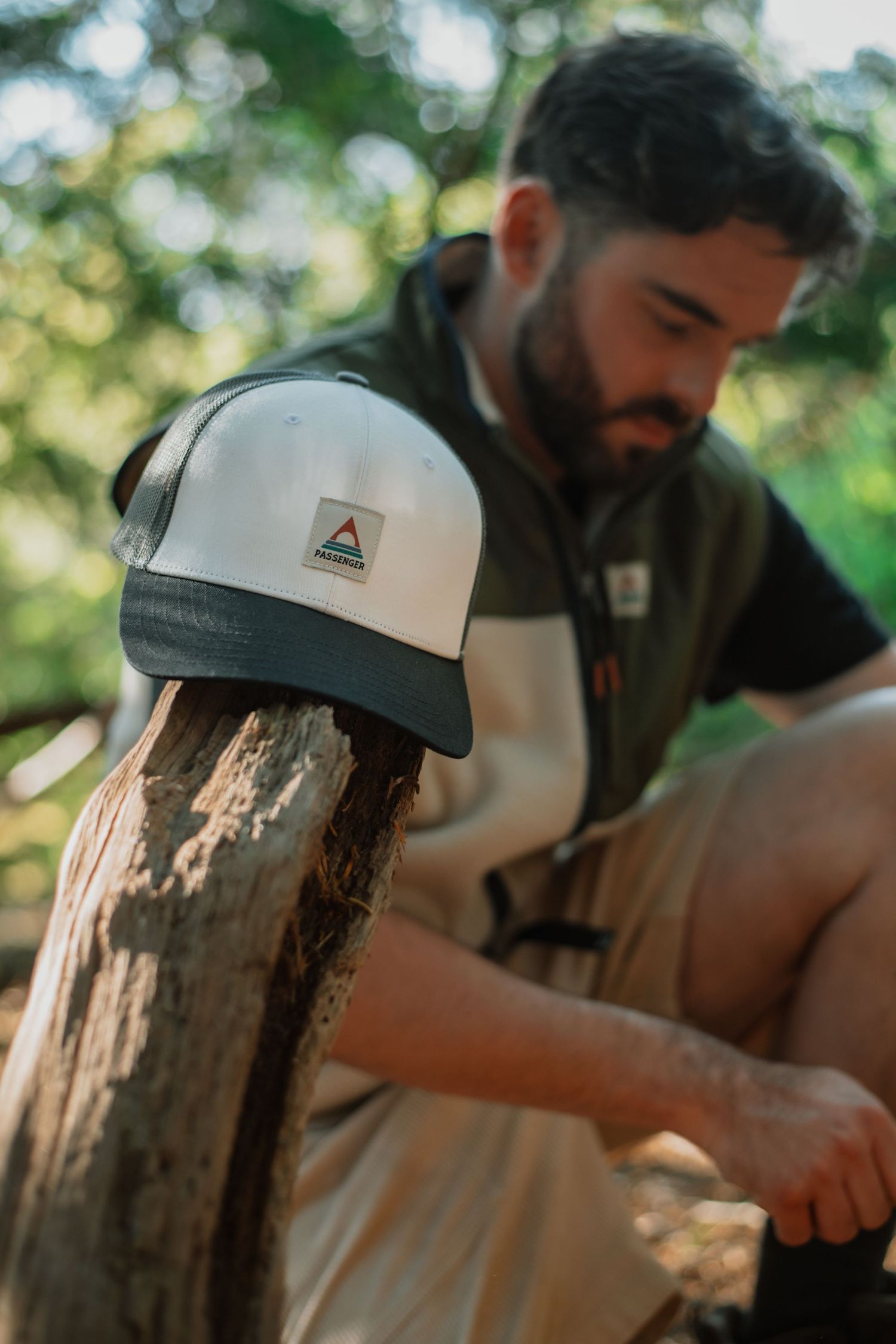 A bearded man in an olive vest sits outdoors, with a white and black Ferrosi cap resting on a wooden post in focus.