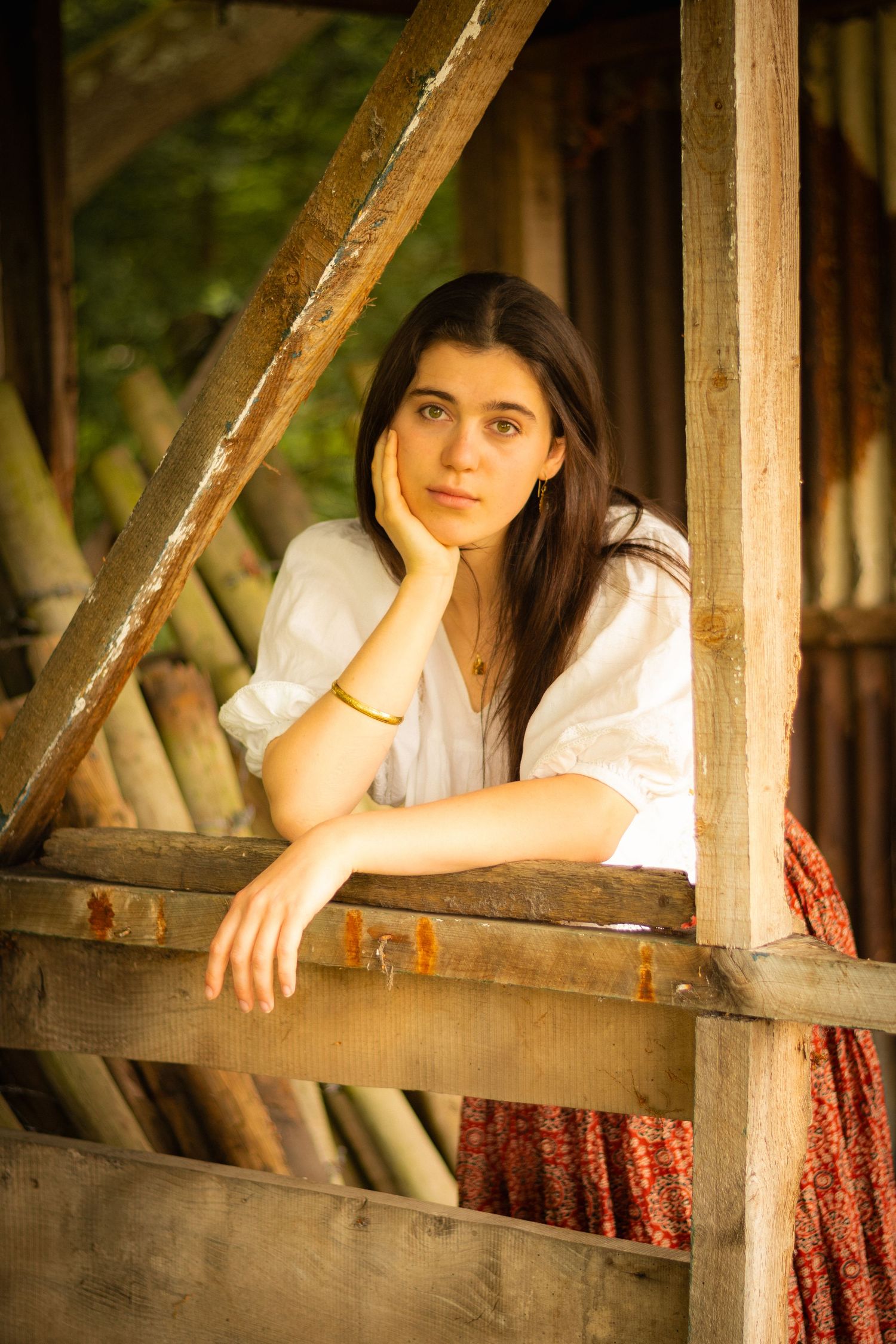 Figure resting contemplatively on wooden balcony railing in white top and patterned skirt.
