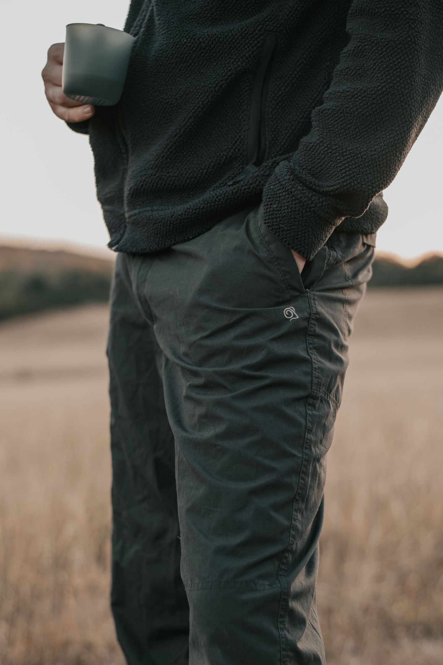Person in dark knit sweater and olive cargo pants holding a green mug outdoors in a golden field.