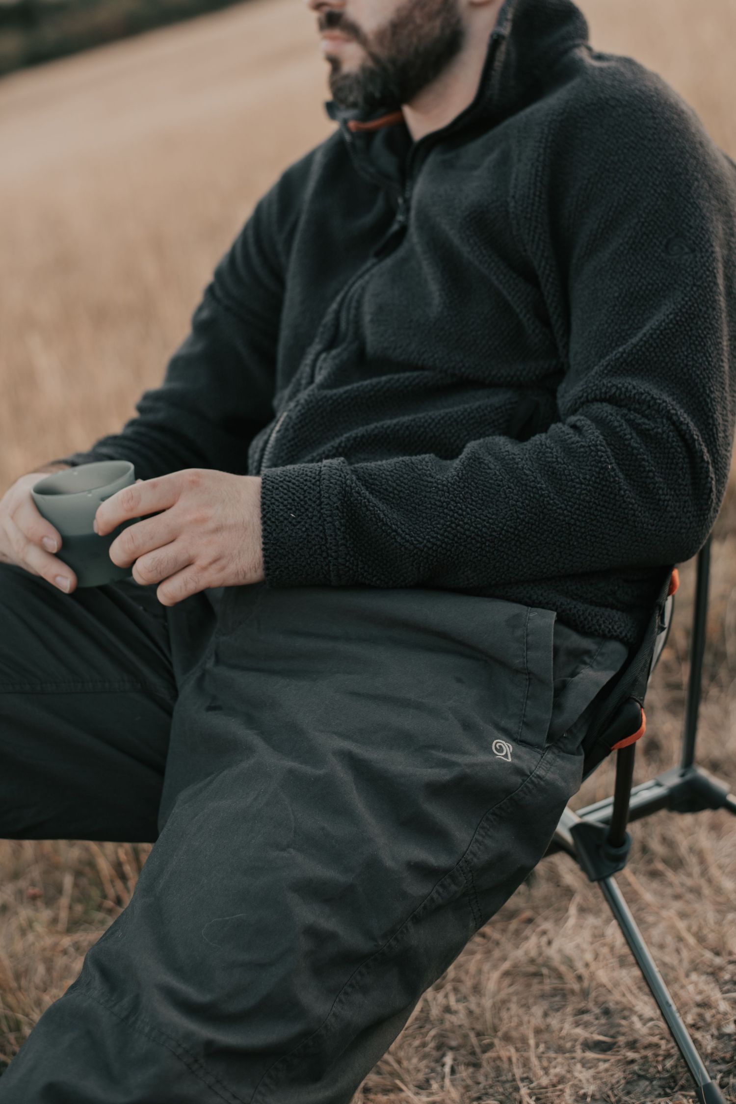 Bearded man sitting in a camping chair outdoors, wearing a dark fleece pullover and cargo pants, holding a mug.