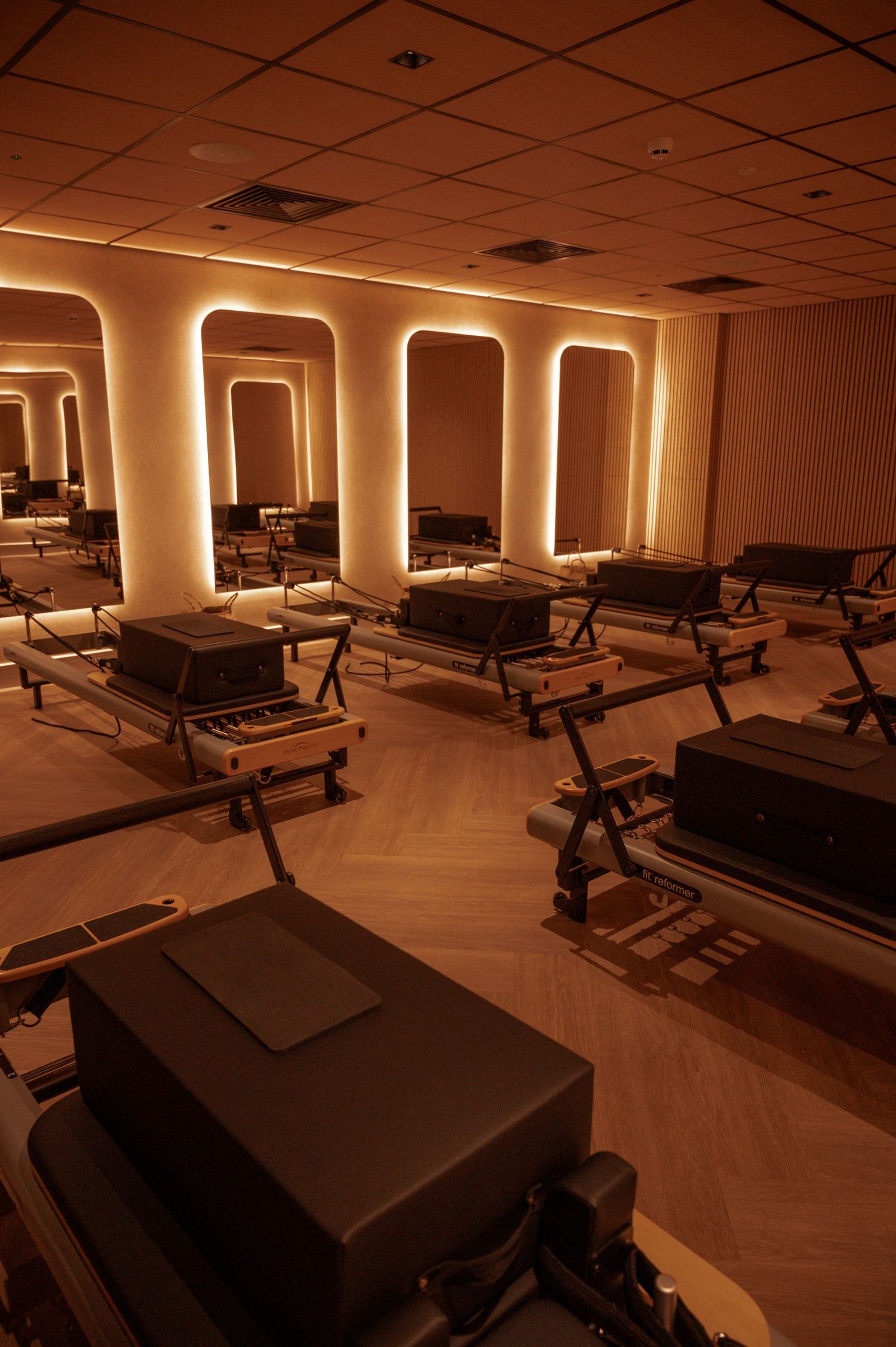 Dimly lit Pilates reformer studio with warm amber lighting, arched wall accents, and rows of reformer machines on herringbone floors.