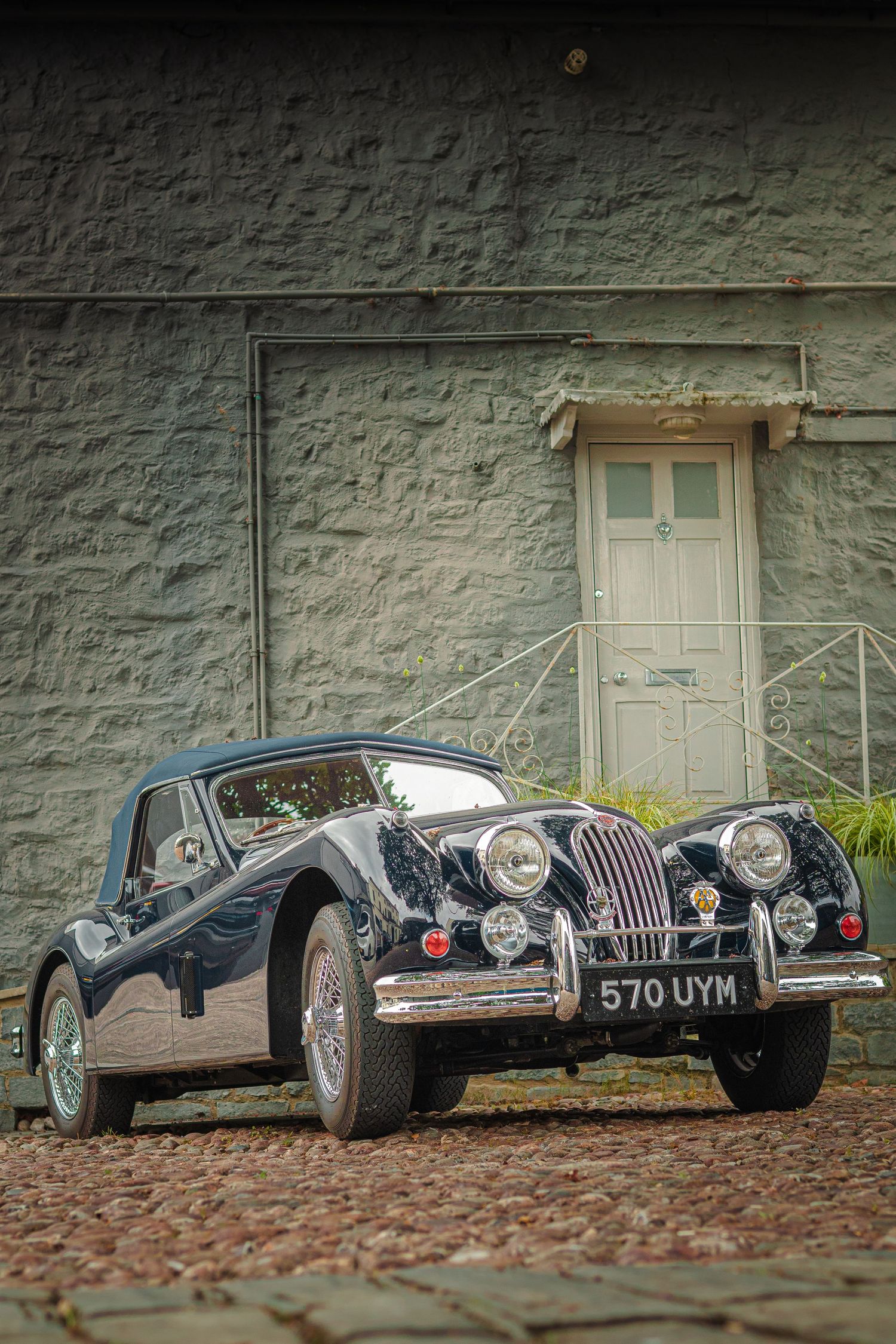 Row of vintage black Jaguar sports cars parked against a stone wall.