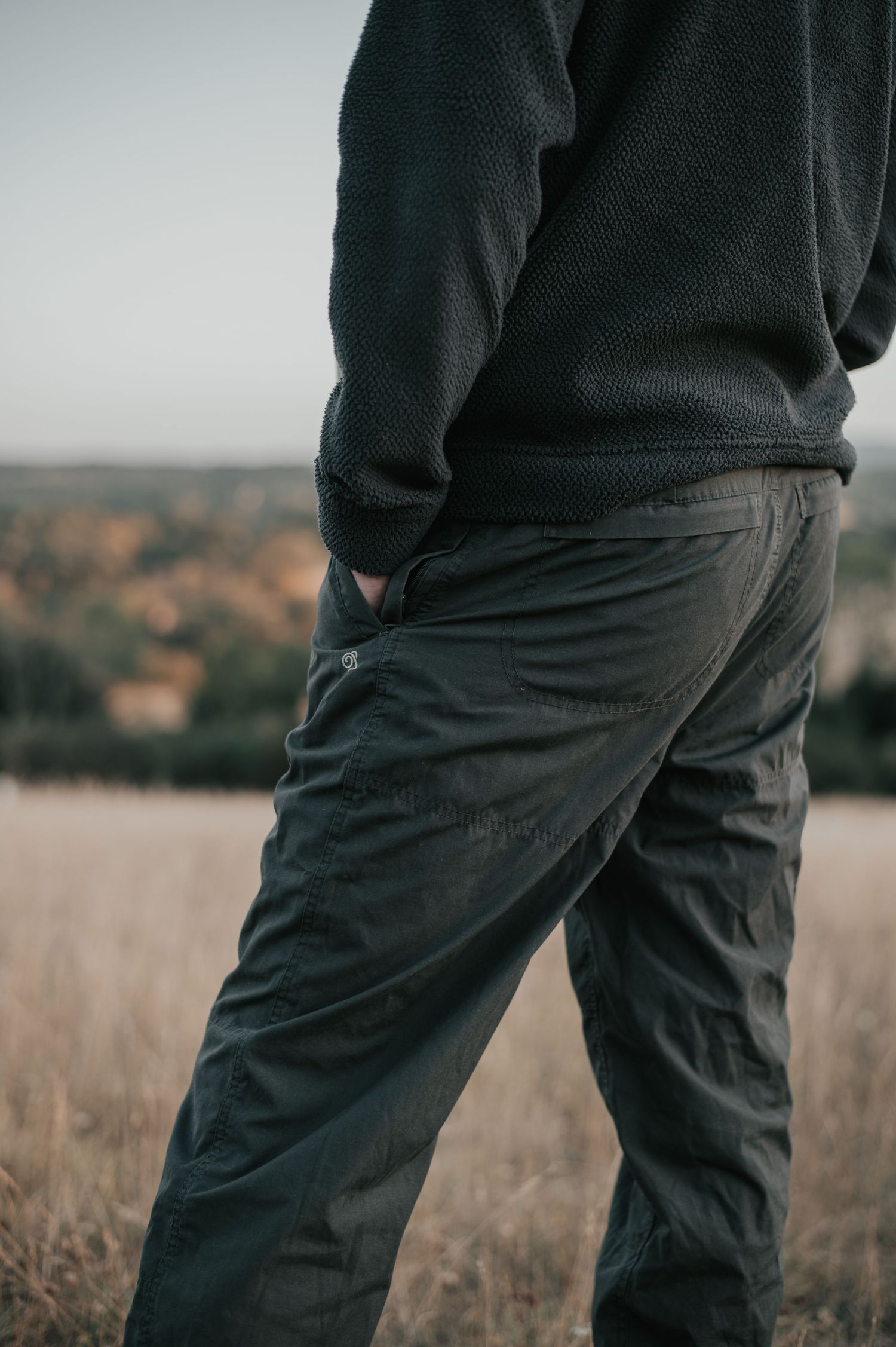 Man wearing dark green cargo pants and sweater standing in a golden field with a scenic landscape backdrop.