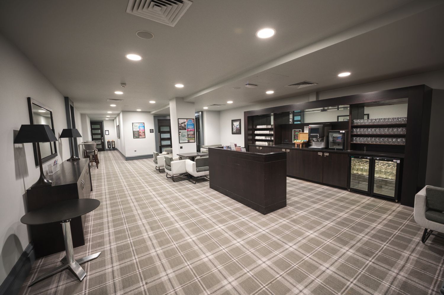 Modern hotel lobby with plaid carpet, reception desk, beverage fridges, and recessed lighting in a sleek gray interior.