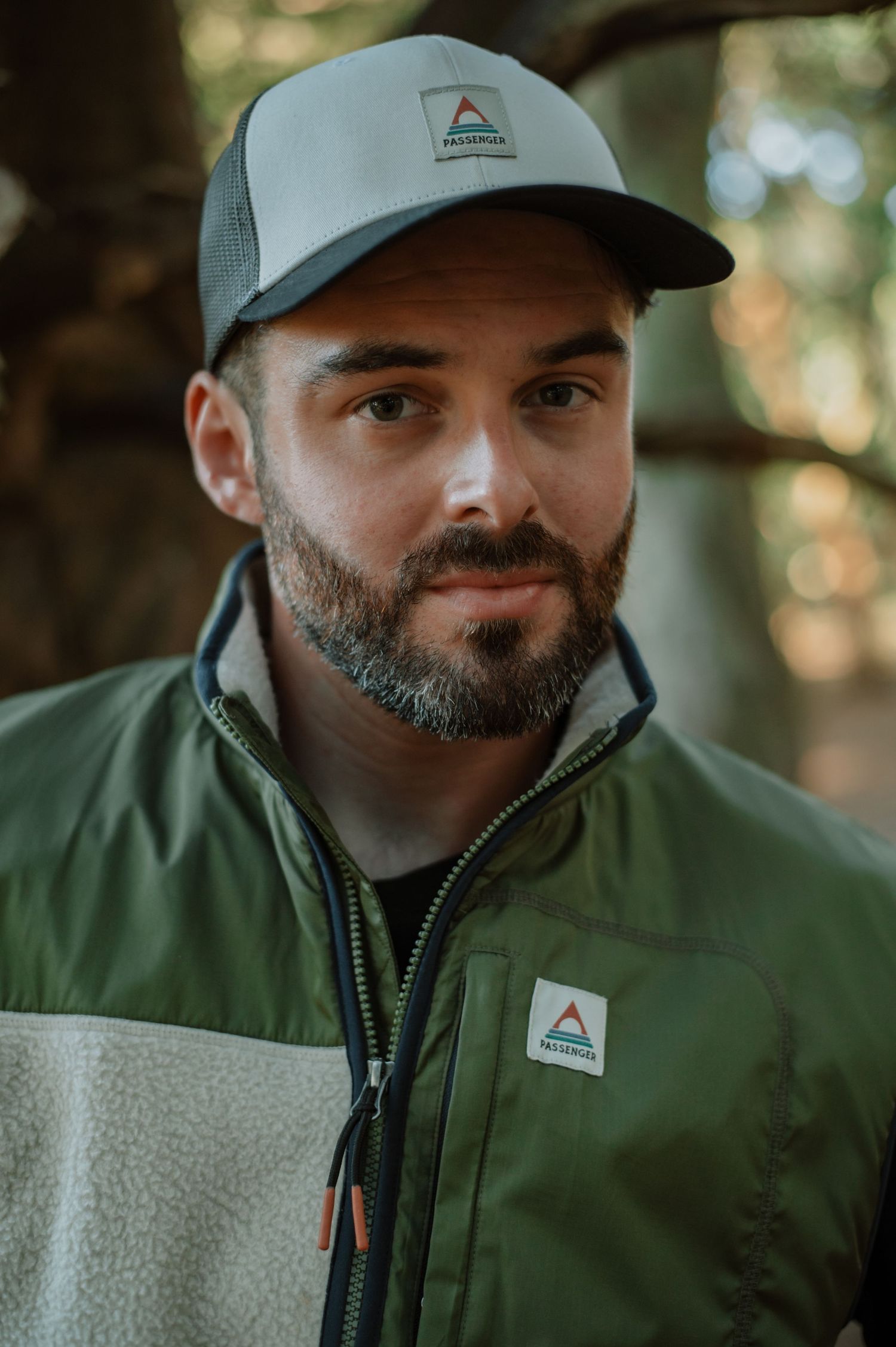 Man with beard wearing green outdoor jacket and matching cap in forest setting.