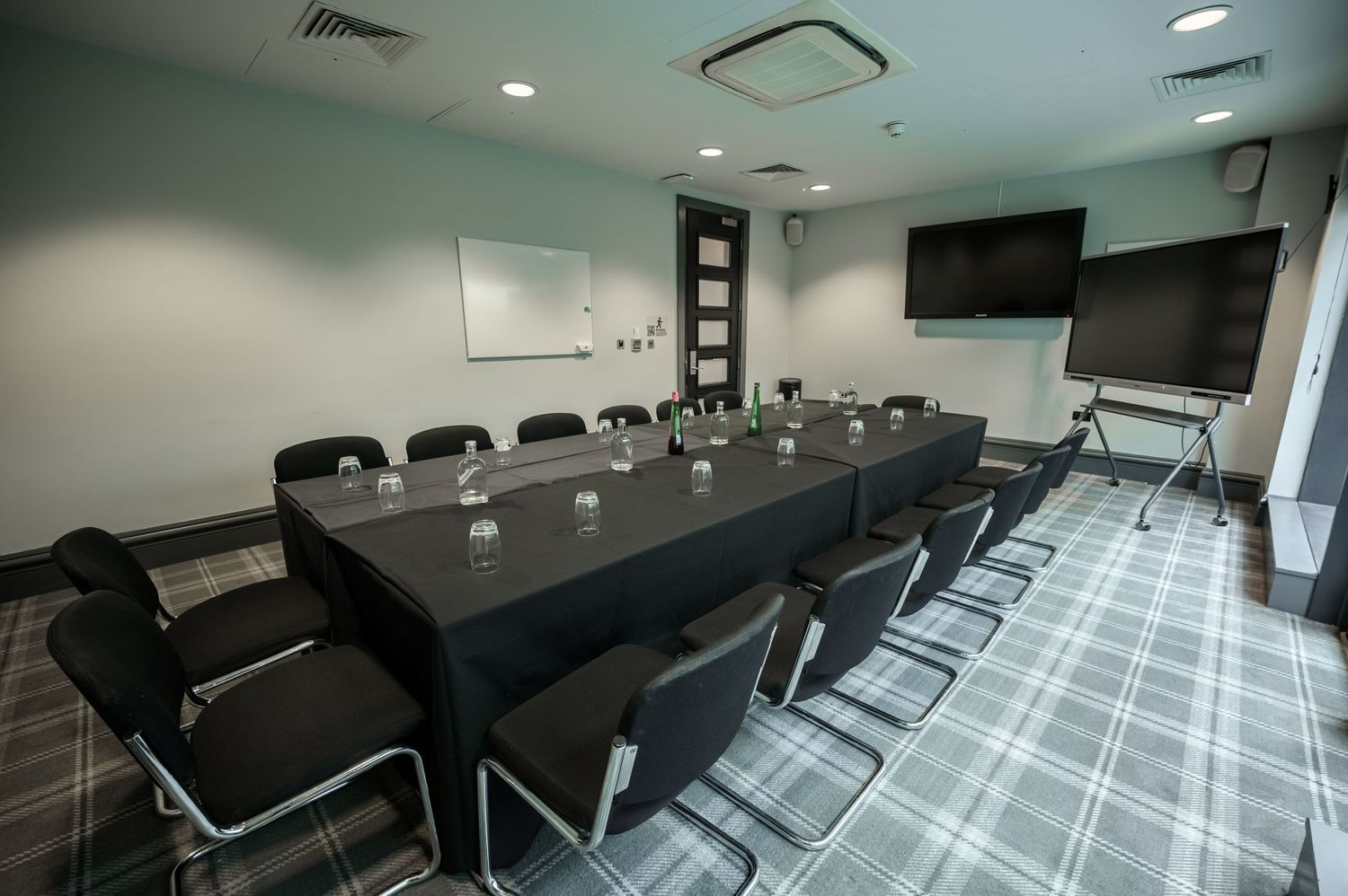 Modern conference room with long black table, ergonomic chairs, whiteboard, and wall-mounted TV on patterned carpet.
