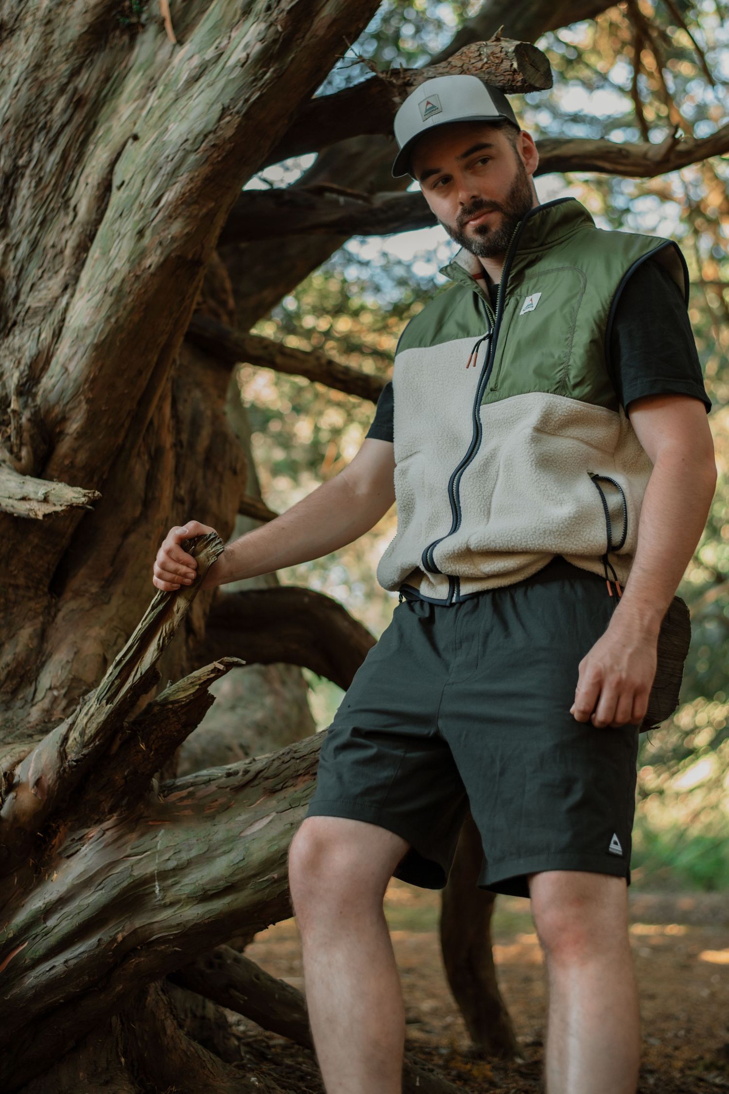Man in green vest, shorts, and cap posing by a large tree in a forest setting.