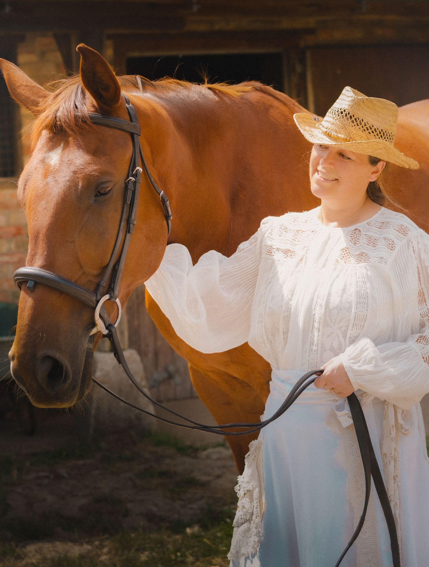 Horse in bridle standing next to someone wearing a white vintage-style dress in a rustic setting.