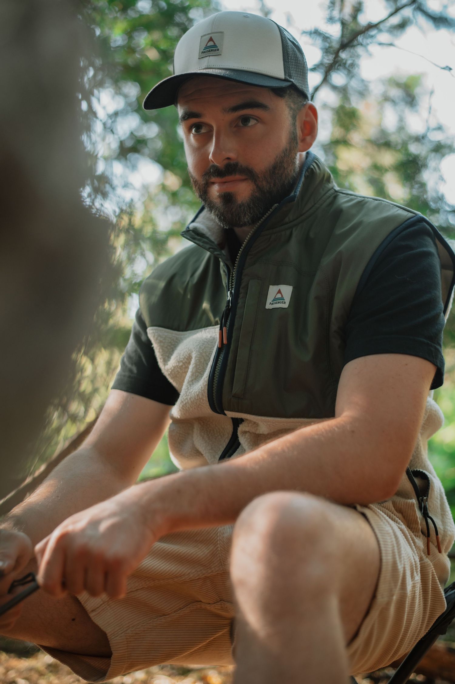 Bearded man in olive vest and trucker hat sitting outdoors in a forest setting.