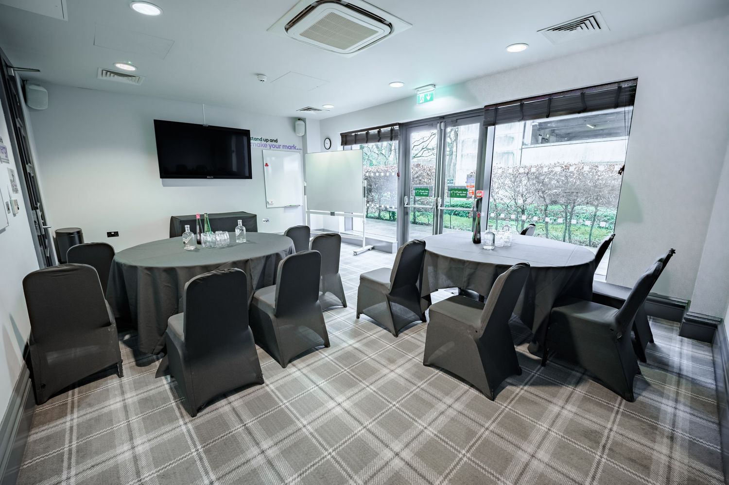 Modern conference room with round tables, black chairs, plaid carpet, wall-mounted TV, and large windows overlooking greenery.