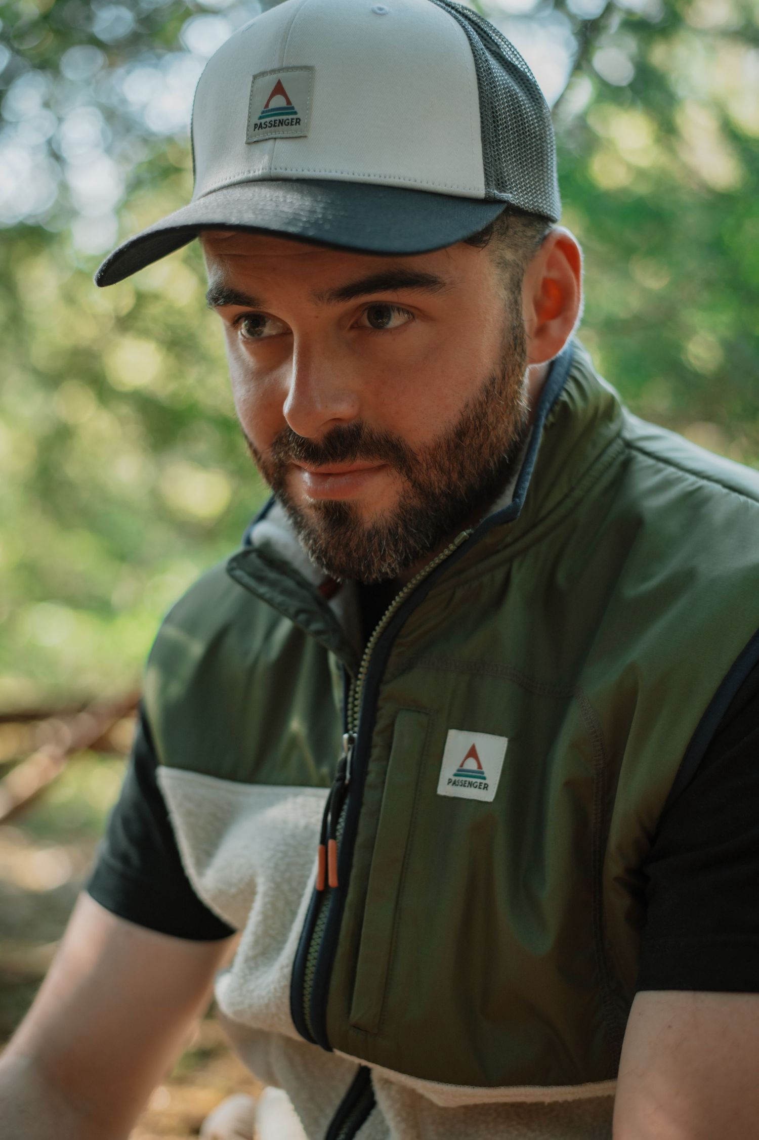 Bearded man wearing a green puffer vest and trucker hat outdoors in a forest setting.