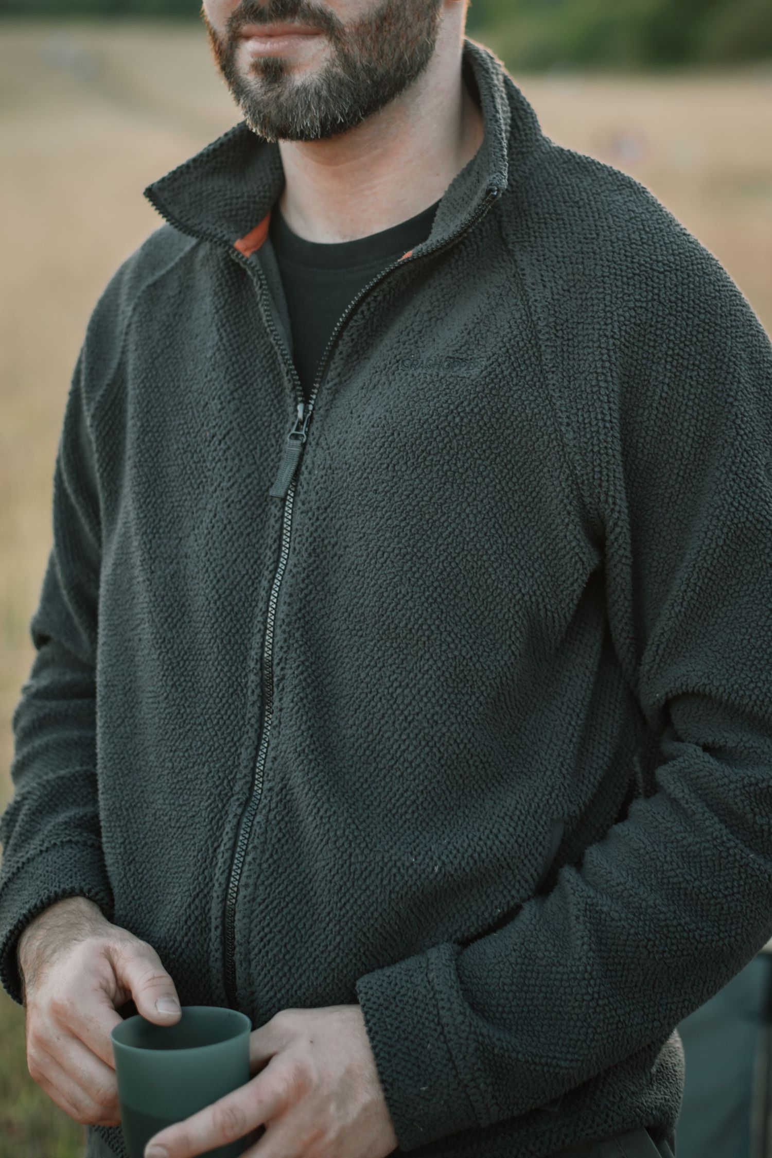 Bearded man wearing a dark green zip-up fleece jacket, holding a matching green mug outdoors.