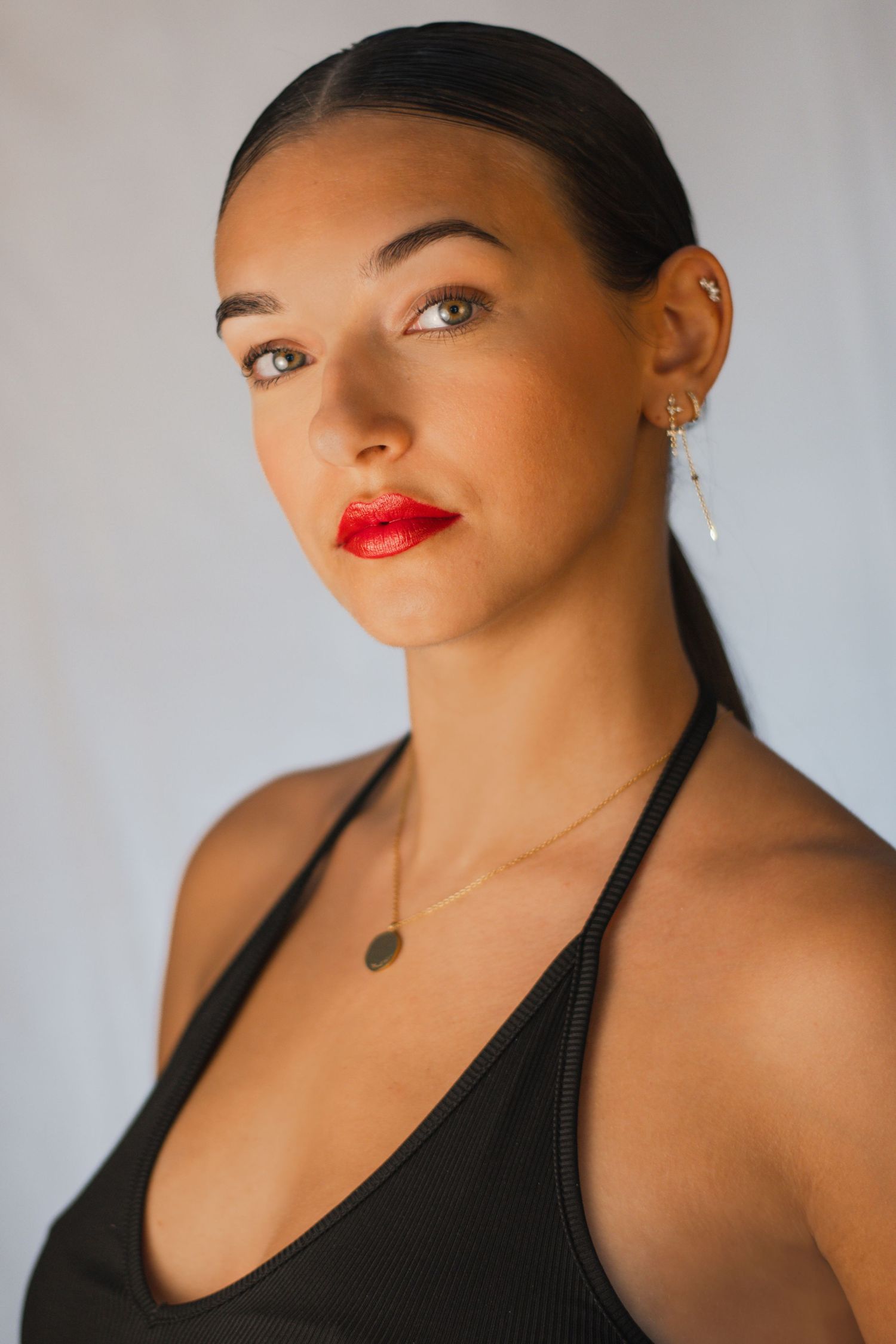 Close-up beauty portrait with bright red lipstick and black tank top against light gray background.