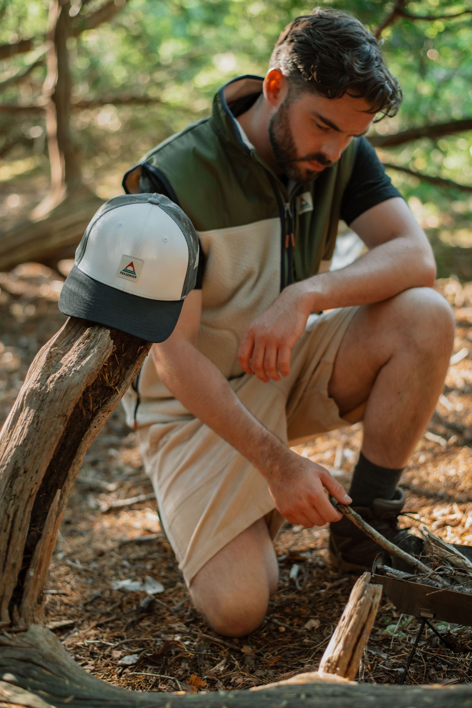 A bearded man in khaki shorts and vest crouches in a forest, starting a campfire with sticks on the ground.