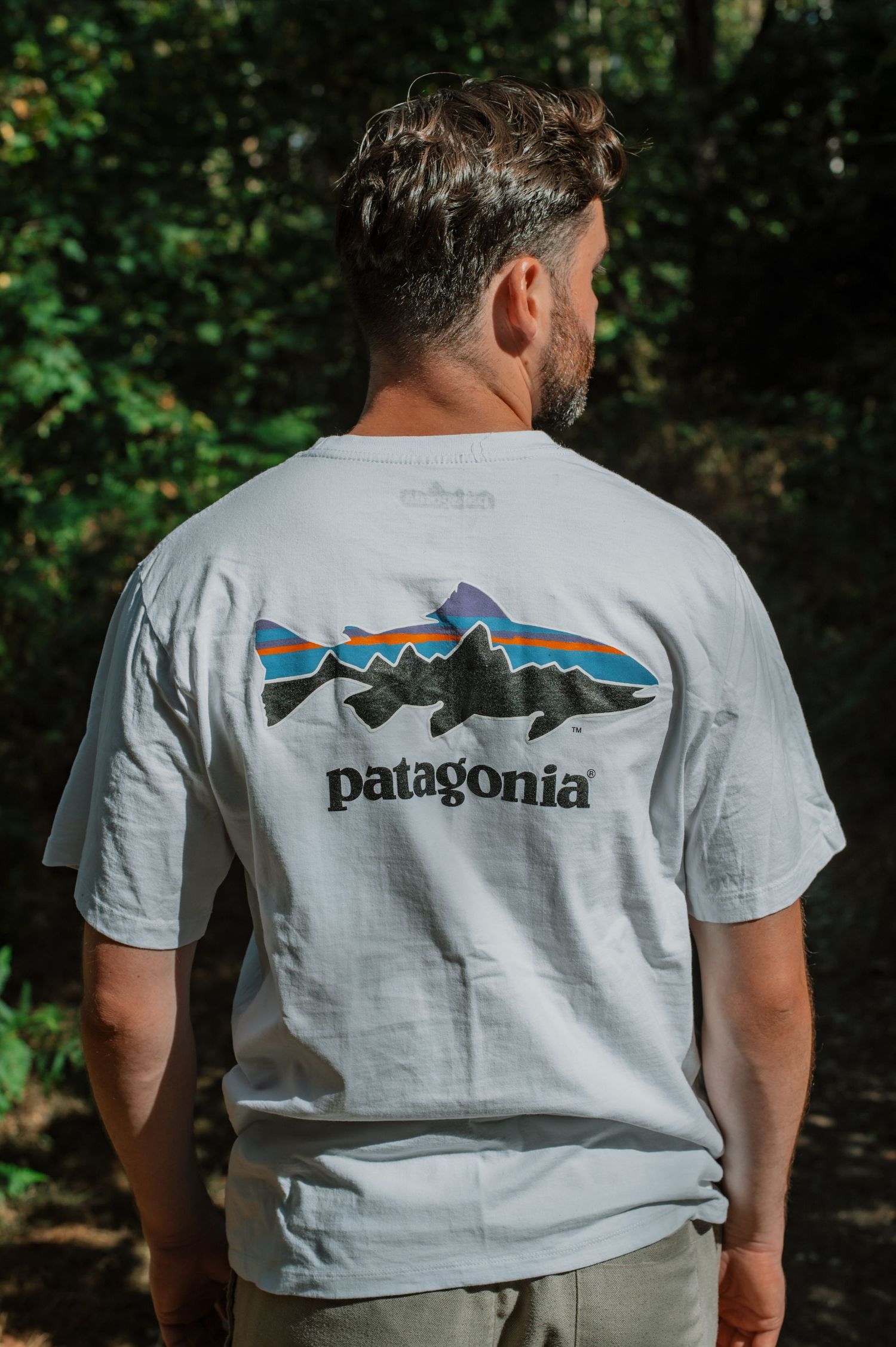 Man wearing a light gray Patagonia graphic t-shirt with mountain and fish logo, standing outdoors in a wooded area.