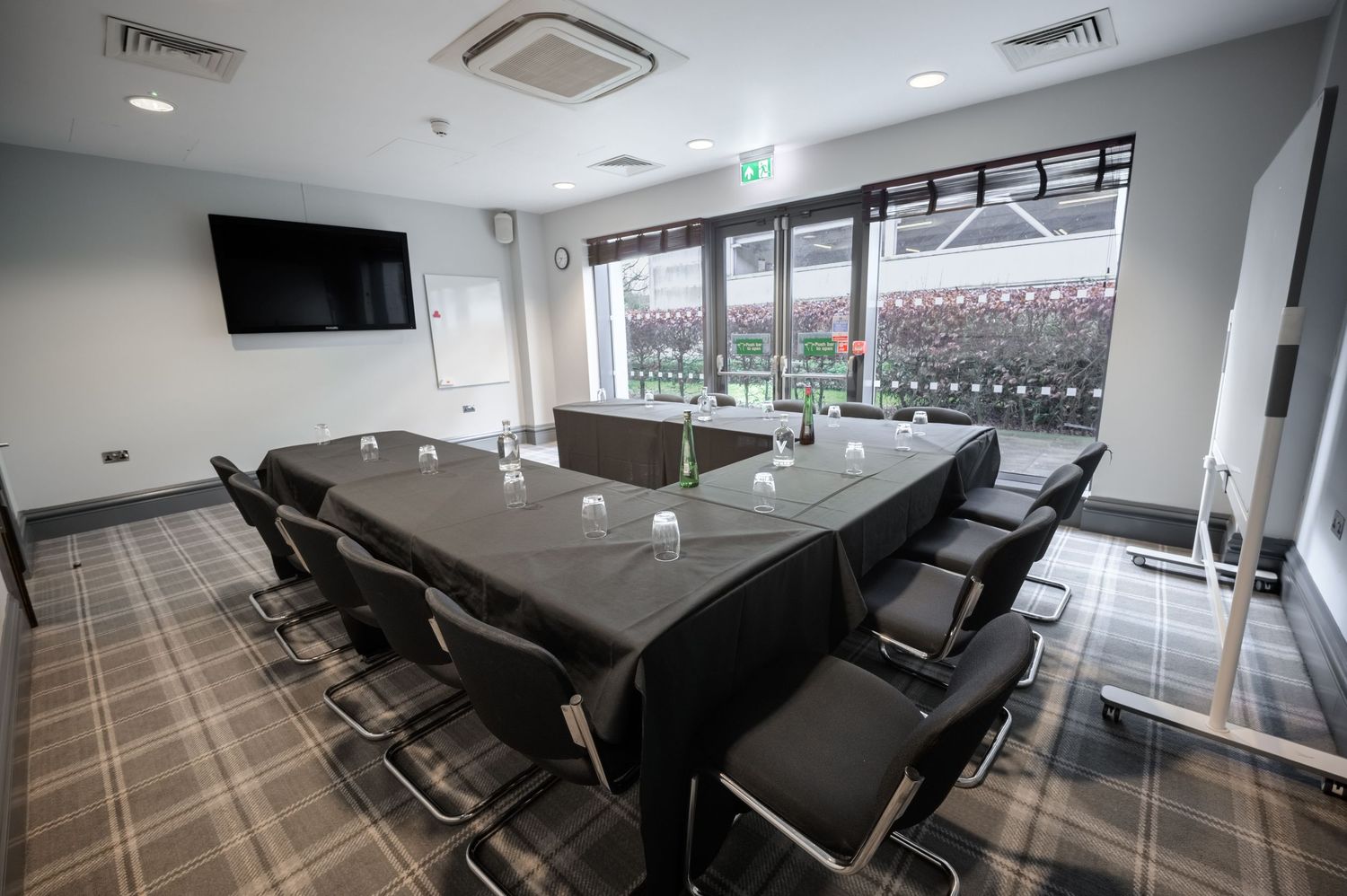 Modern boardroom with U-shaped table, black leather chairs, wall-mounted TV, whiteboard, and large windows overlooking a stadium.