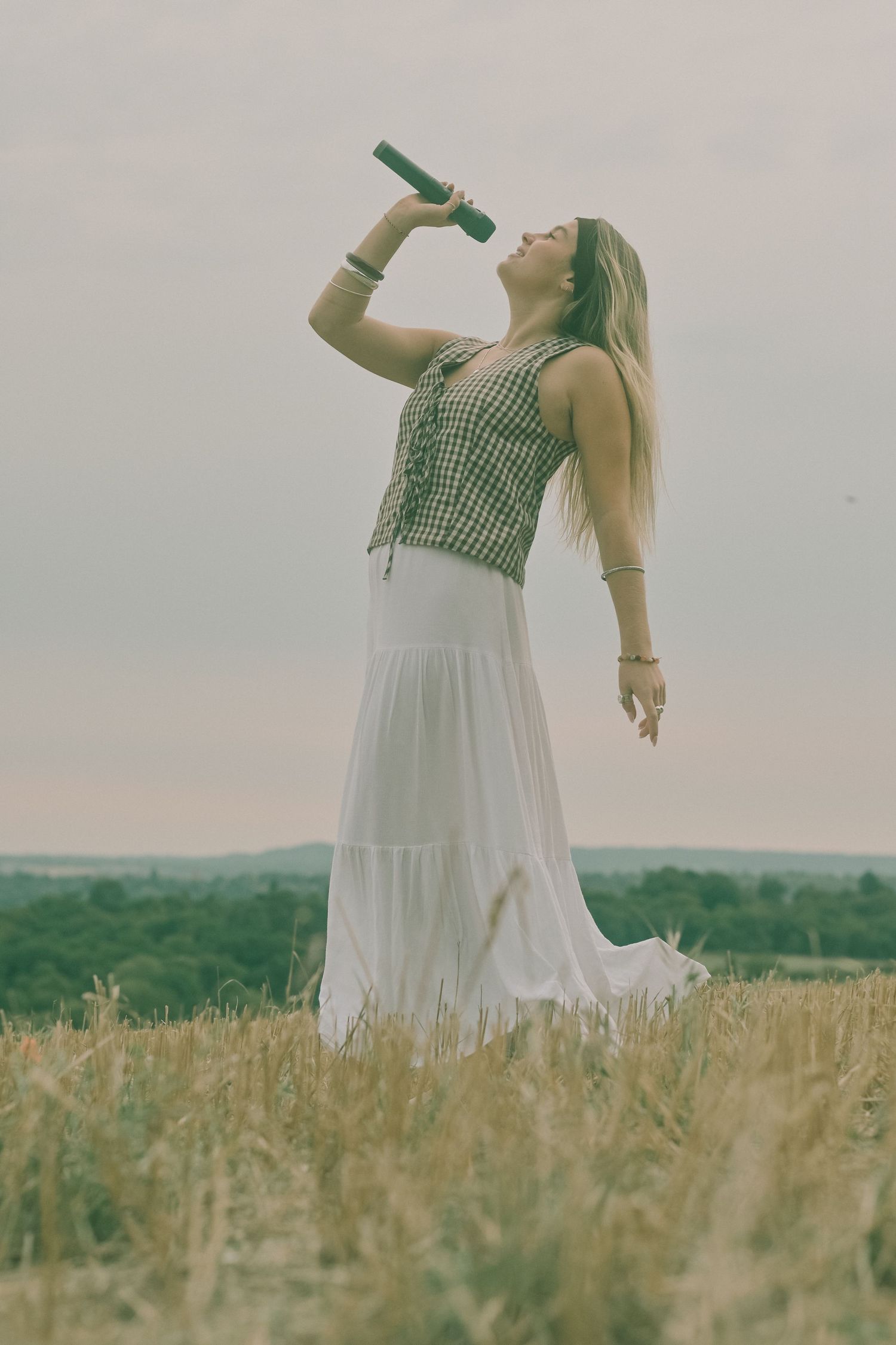 Figure in gingham and white dress dances with bottle in golden field at sunset.