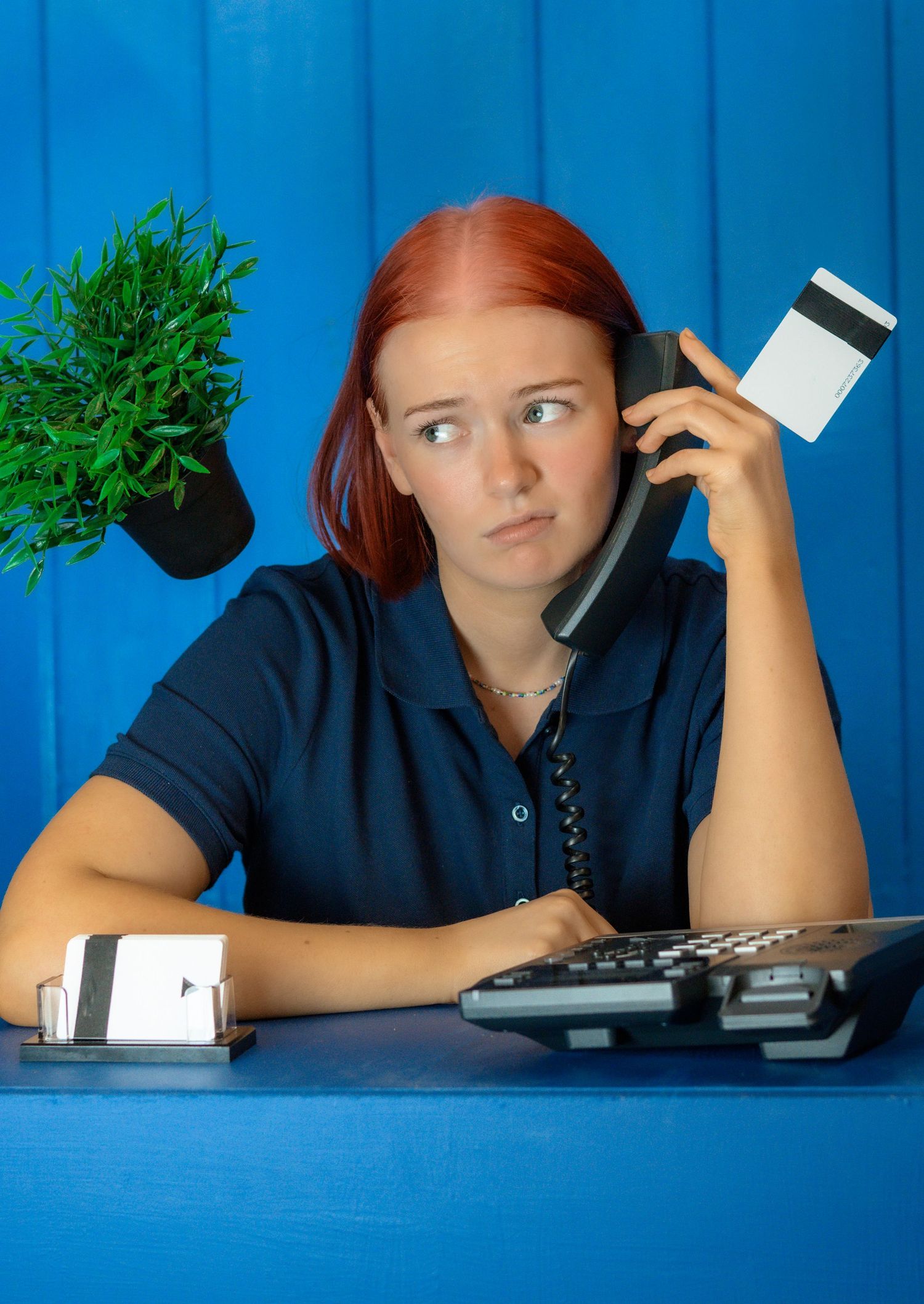 Redheaded office worker looking frustrated while holding phone with plant on desk.