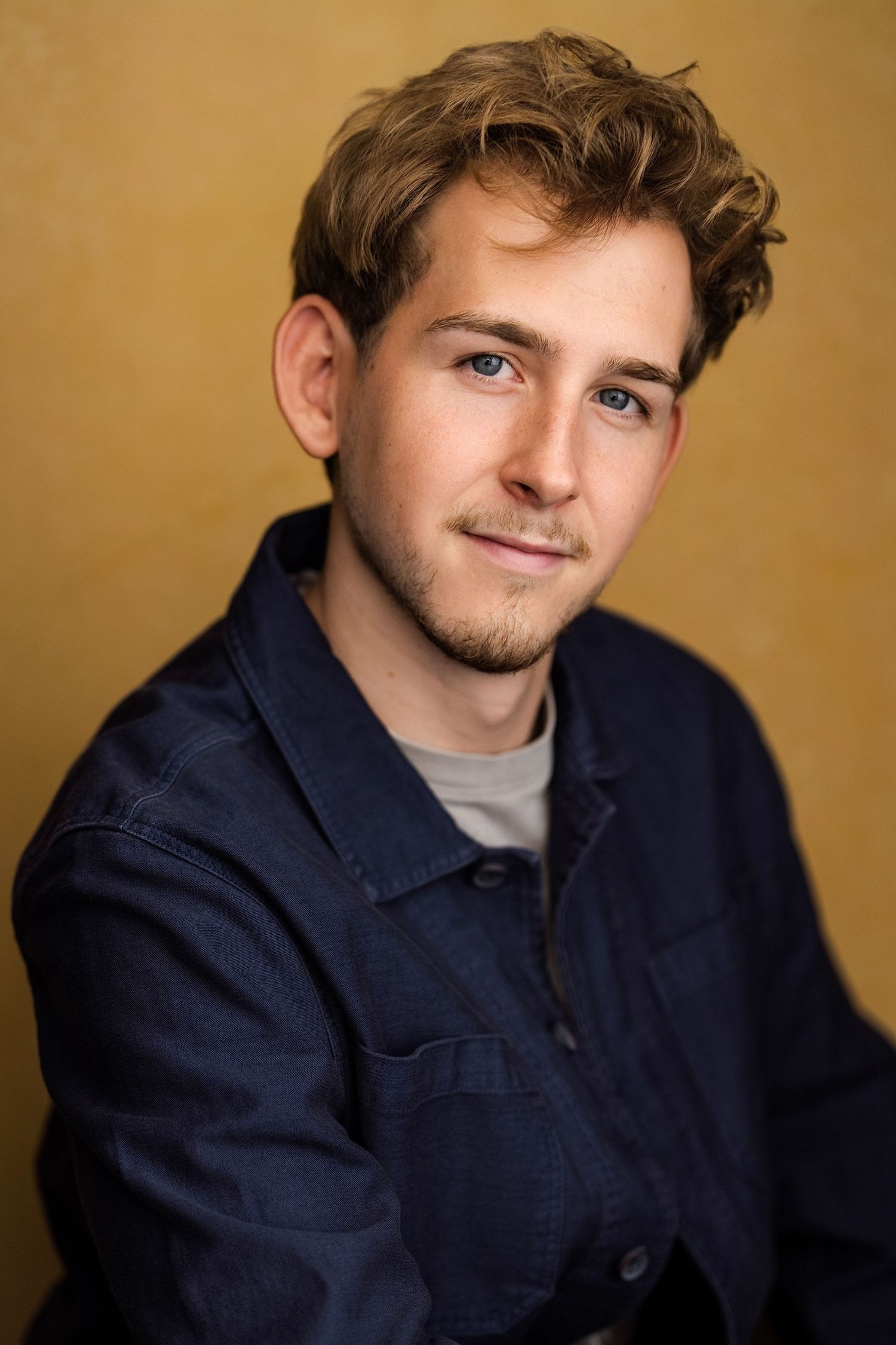 Young man with blue eyes and wavy blonde hair wearing a navy shirt, posing for a professional headshot portrait.