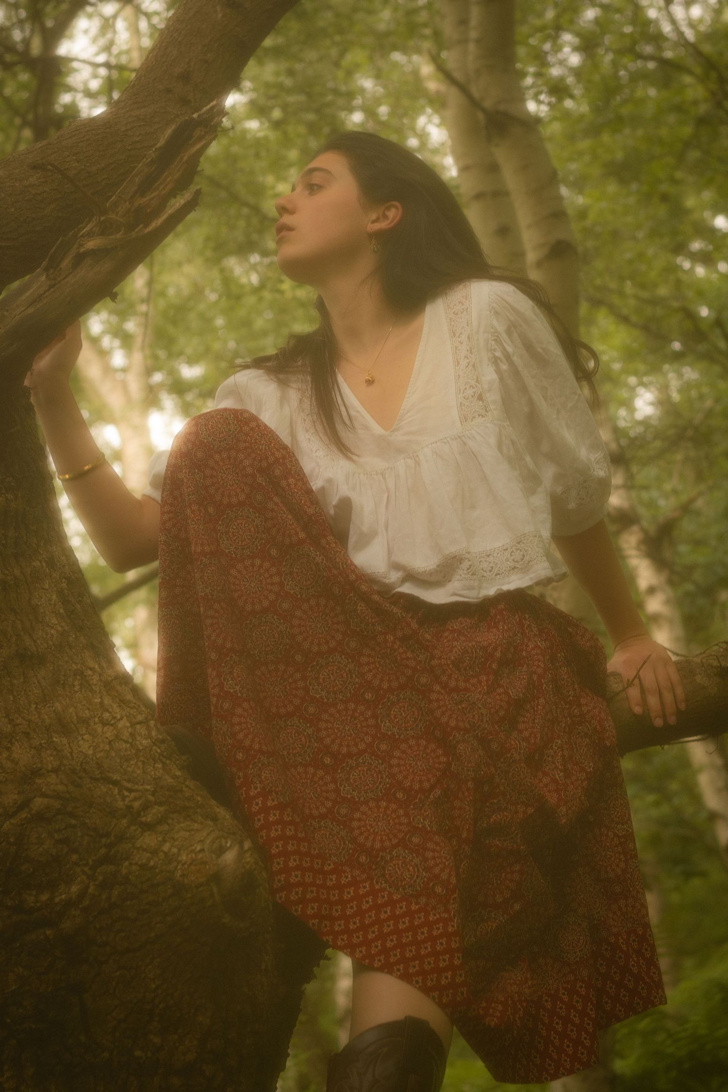 A dreamy vintage-style photo series of someone in a red knit shawl and white dress climbing a tree in dappled sunlight.