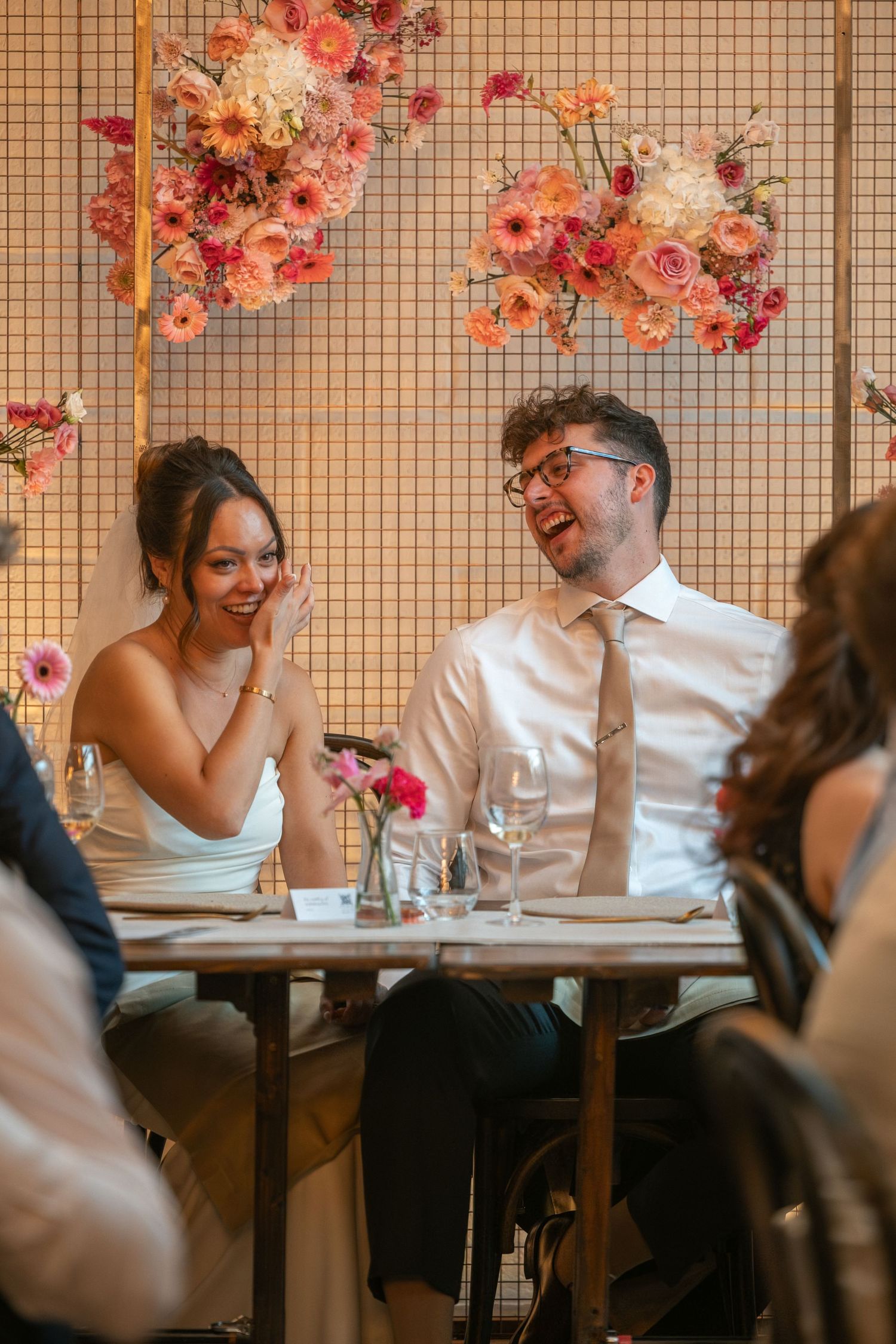 Bride and groom laughing joyfully at wedding reception table adorned with pink floral arrangements.
