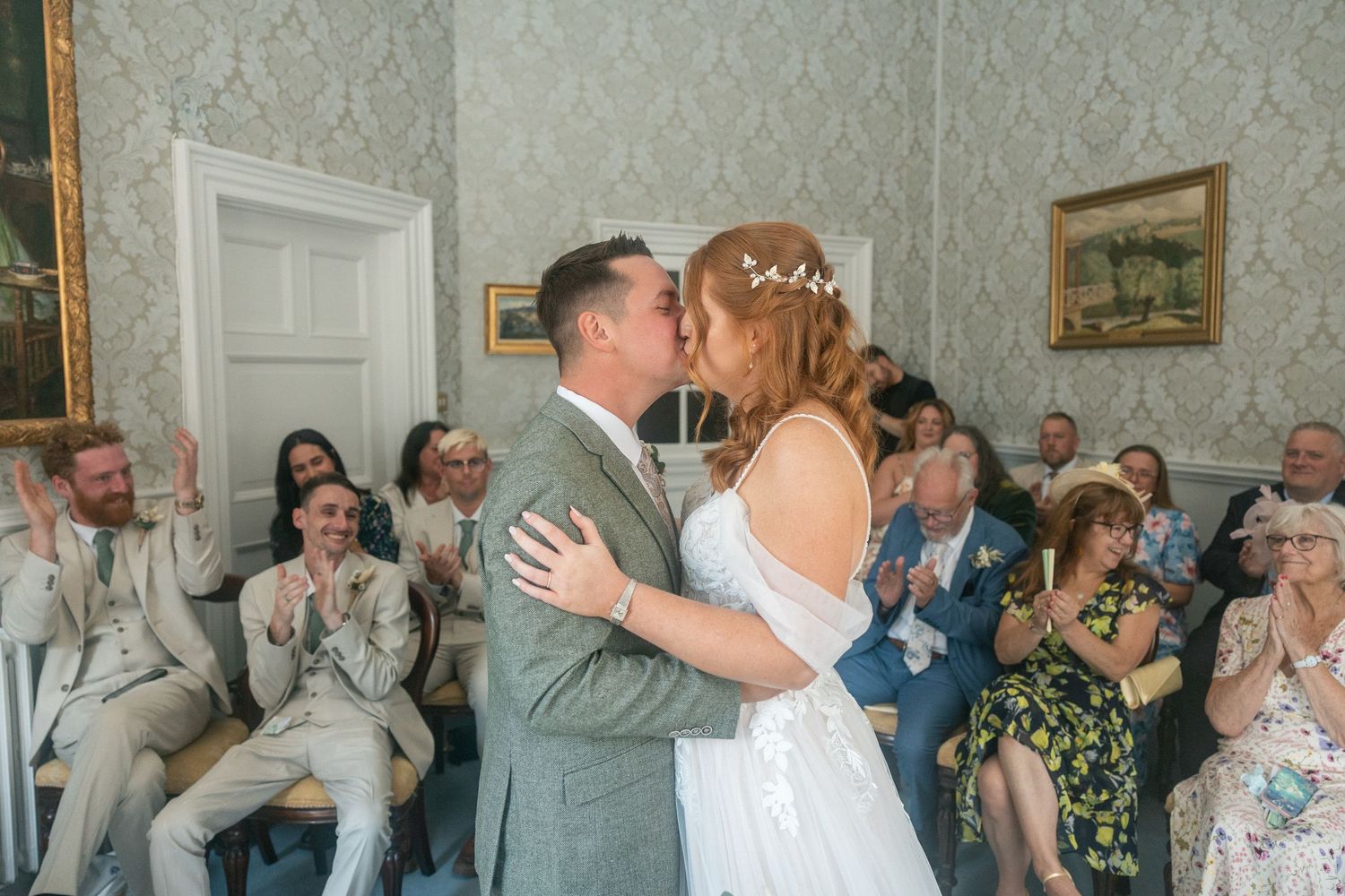 Bride and groom share first kiss at wedding ceremony while guests applaud in The Guildhall Bath.