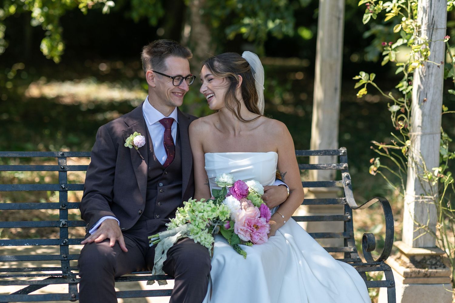 A bride and groom sit on a park bench smiling at each other, bride holding a pink and green bouquet.