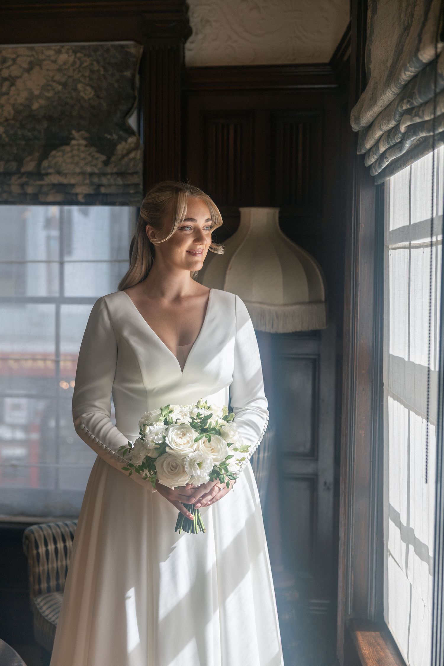 Bride in elegant white long-sleeve wedding gown holding white rose bouquet, standing by sunlit window in classic interior.
