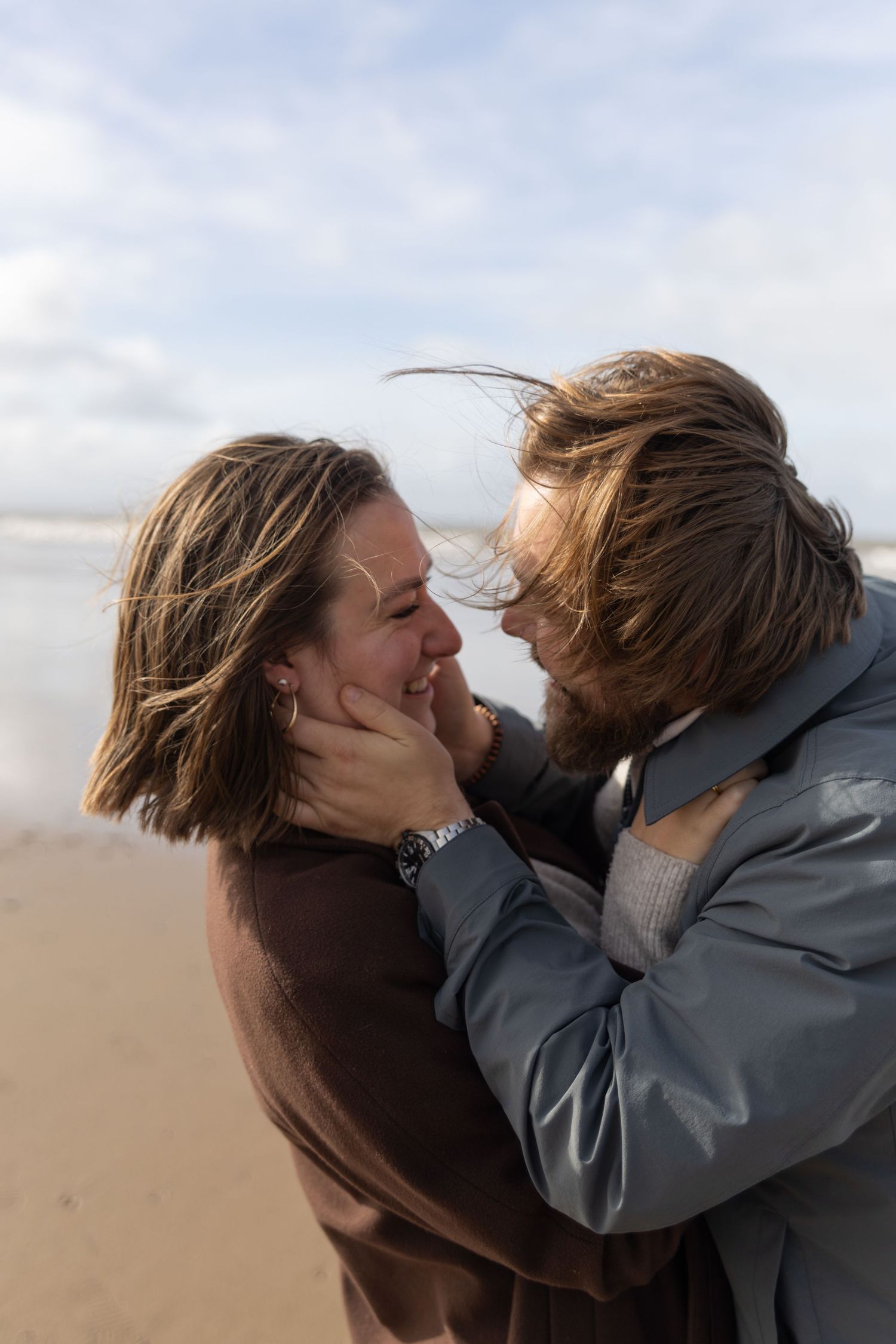 Two people share an intimate embrace with wind-blown hair on a sunny beach.