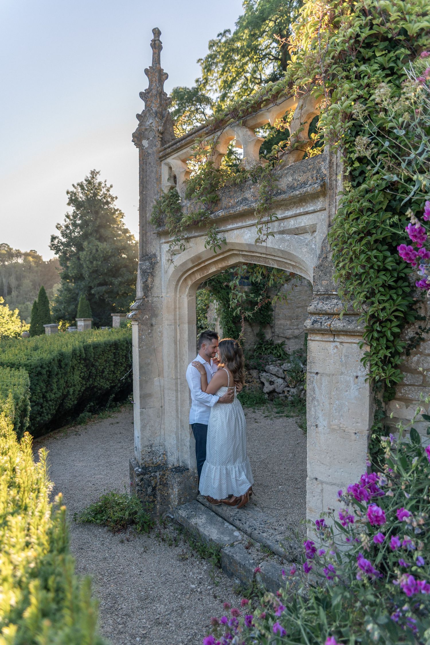 Couple embracing and kissing under a stone arch in a lush garden with purple flowers and ivy at golden hour.