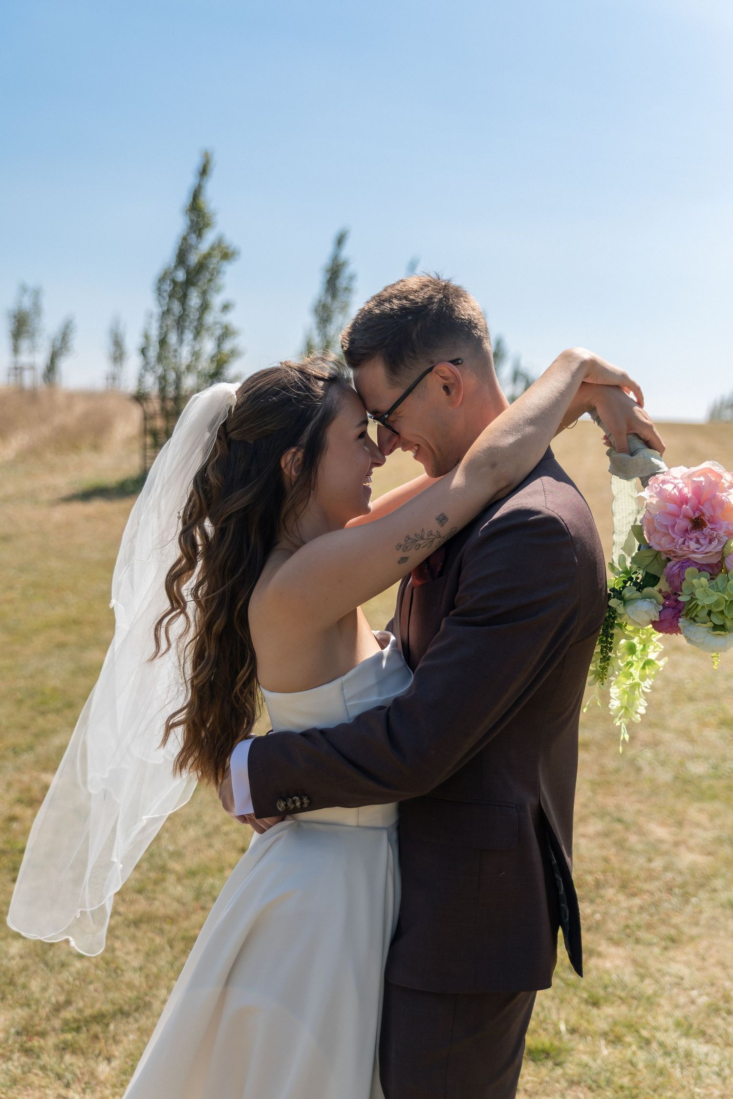 Bride and groom embracing outdoors on sunny wedding day, bride holding pink bouquet wearing veil, groom in dark suit.