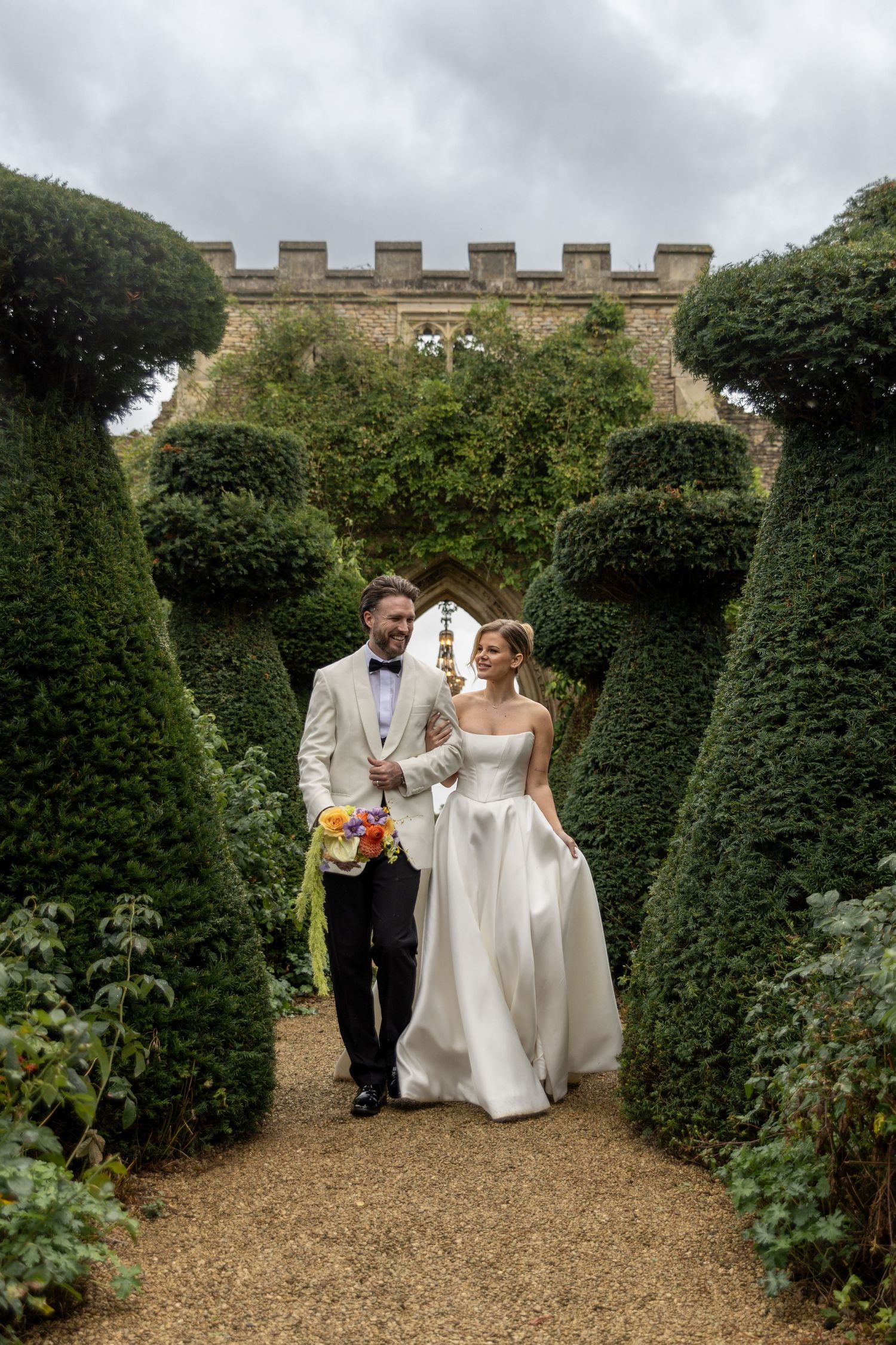 A wedding couple walks through a manicured garden path with topiary and stone castle walls.