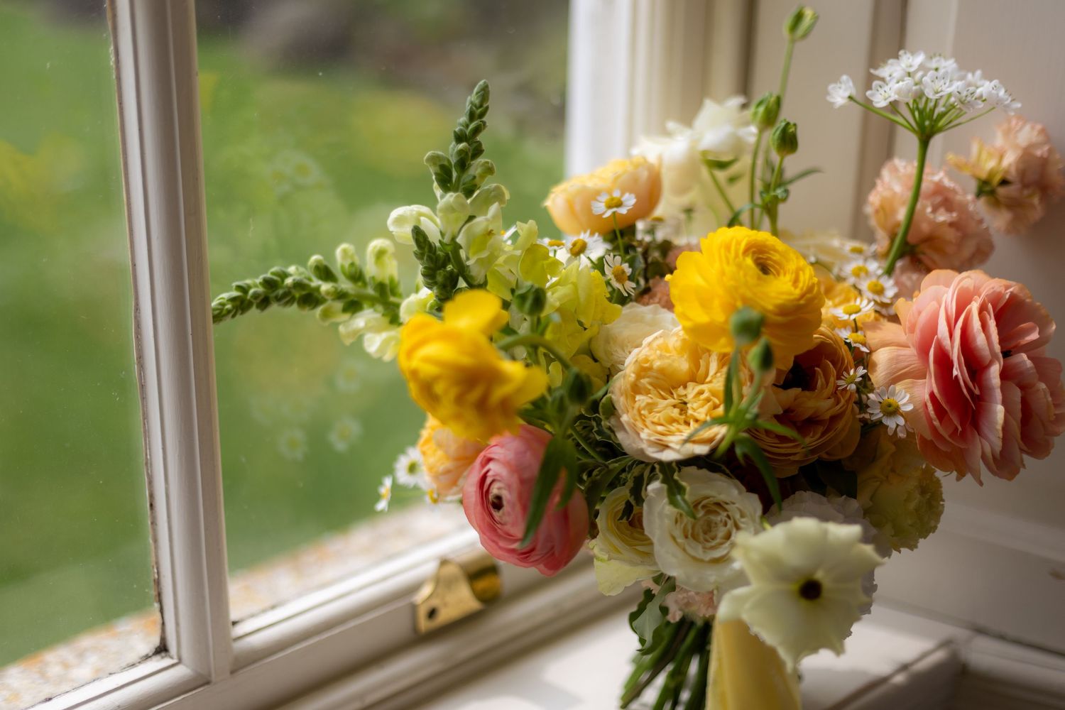 Colorful spring wedding bouquet with yellow, pink, and white flowers displayed on a white windowsill in natural light.