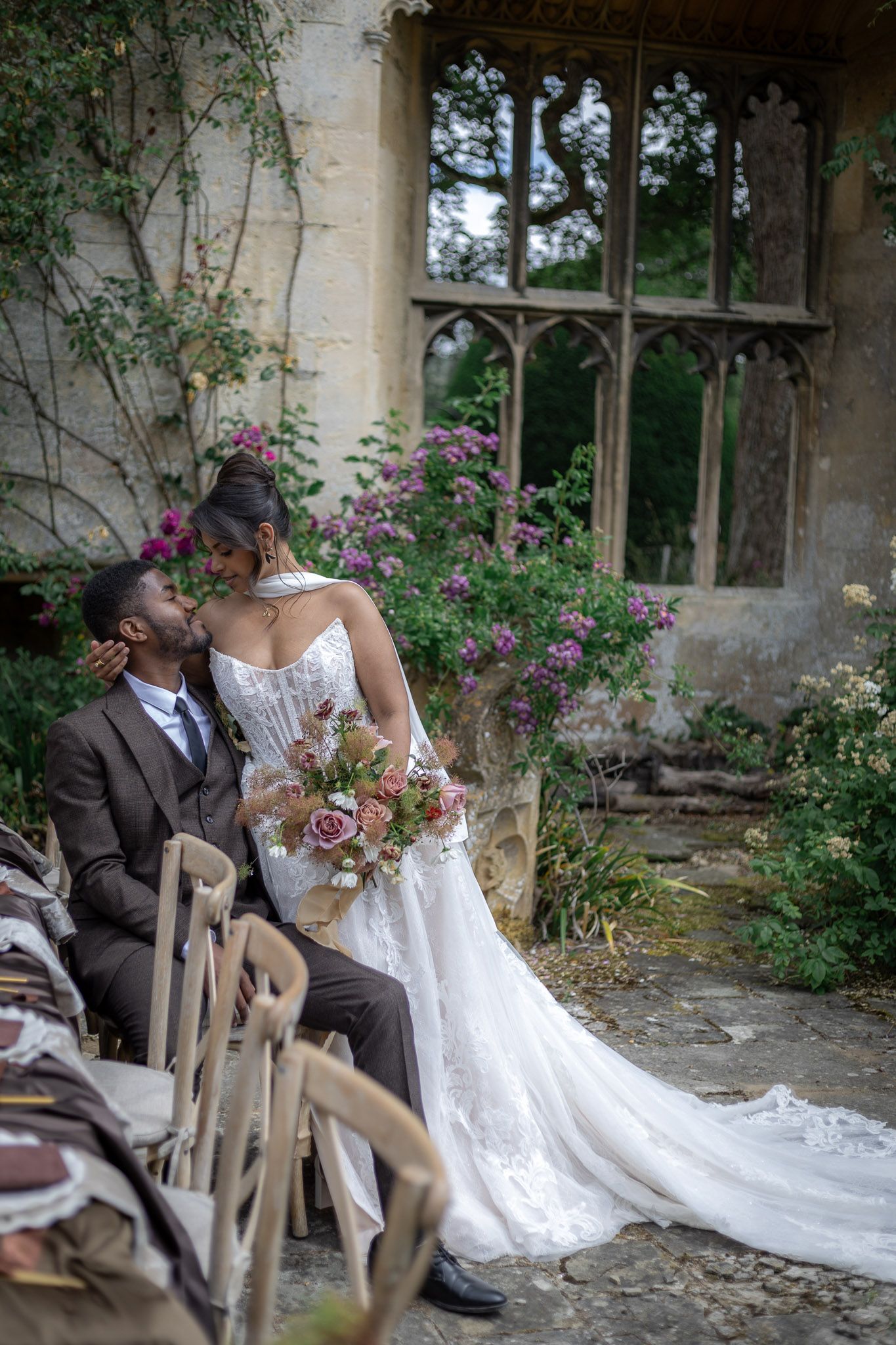 A bride and groom at Sudeley Castle share a romantic kiss at an outdoor wedding ceremony surrounded by lush greenery and blooming flowers.
