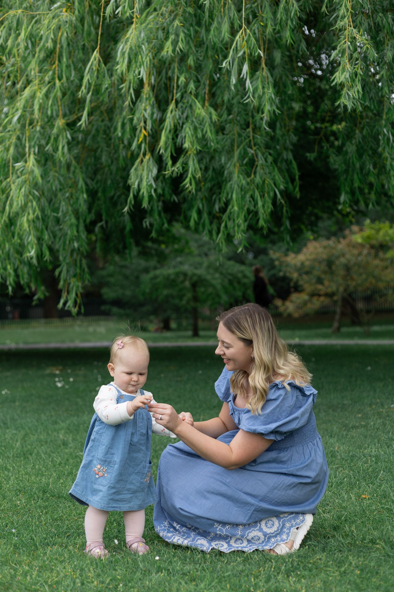 Mother and child share playful moment on grass under willow trees.