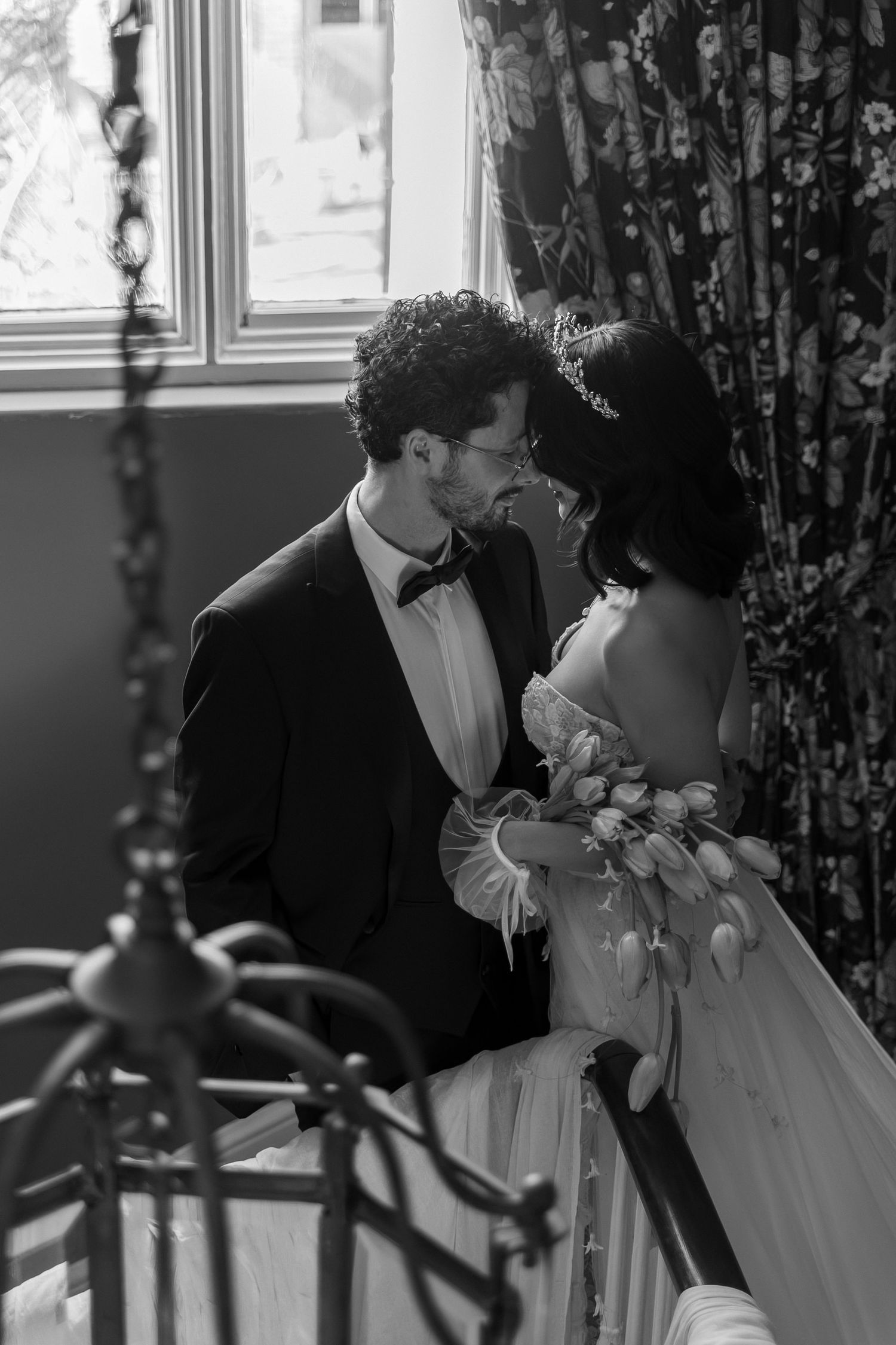 A romantic black and white wedding photo of a couple in formal attire sharing an intimate moment by a window.