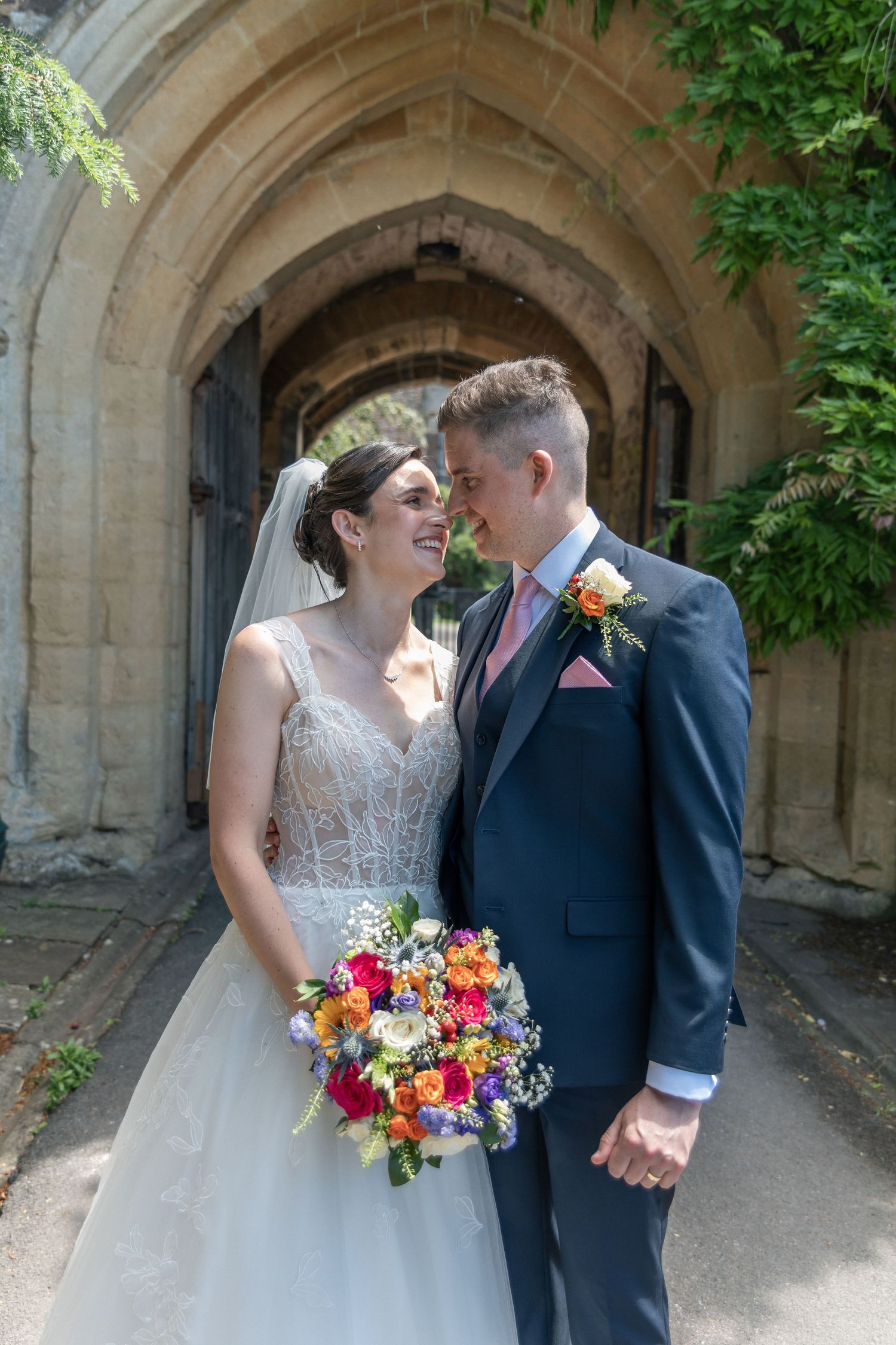 Bride and groom smiling at each other outside a stone church arch, bride holding colorful bouquet, groom in navy suit.