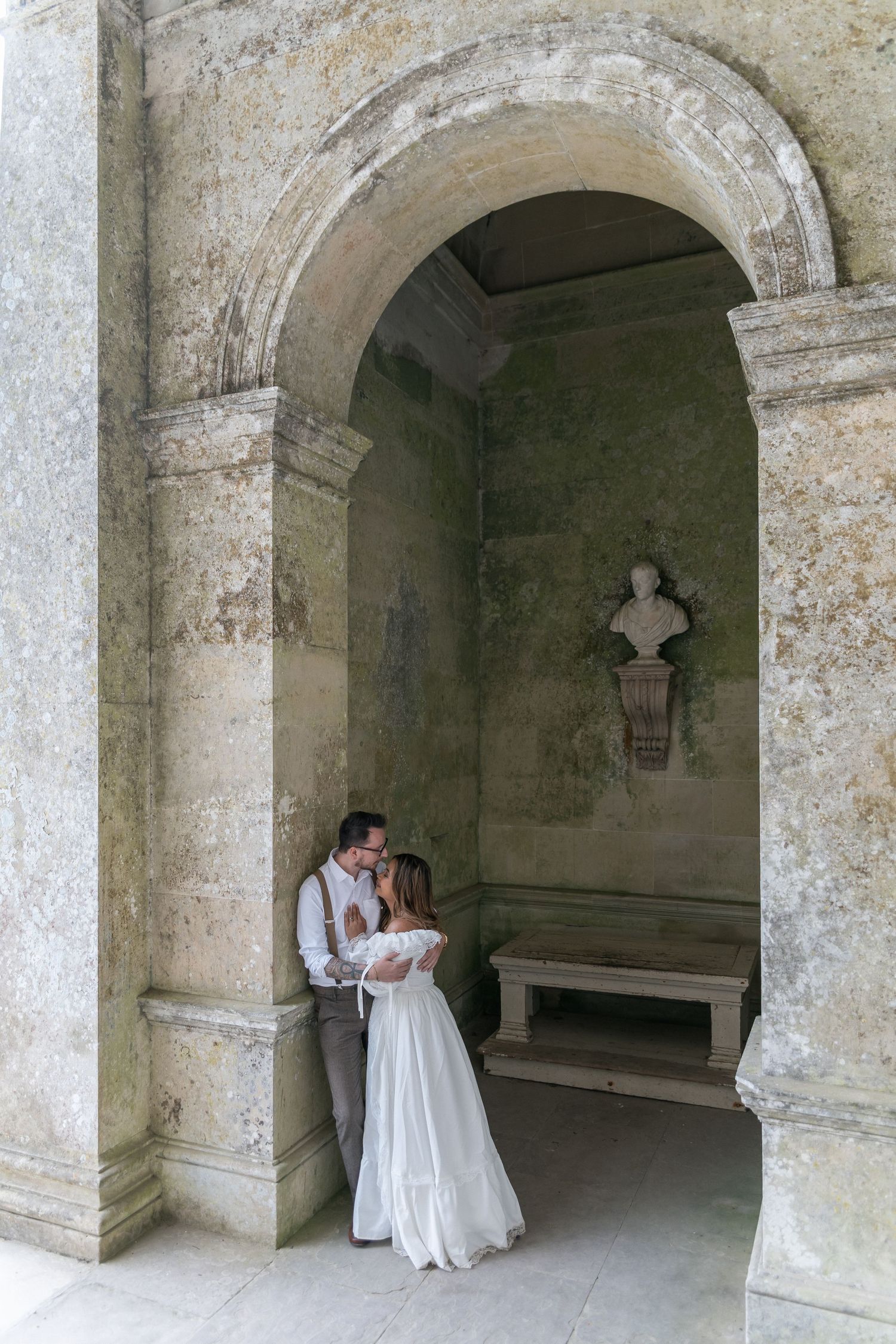 Bride and groom embrace under a stone arch in an ancient building with a classical bust sculpture in the background.