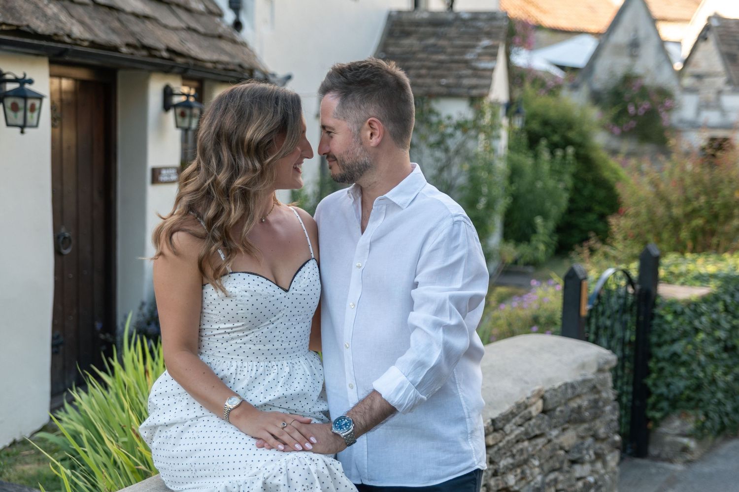 Couple in white outfits share a romantic moment outside a charming cottage, gazing lovingly at each other.