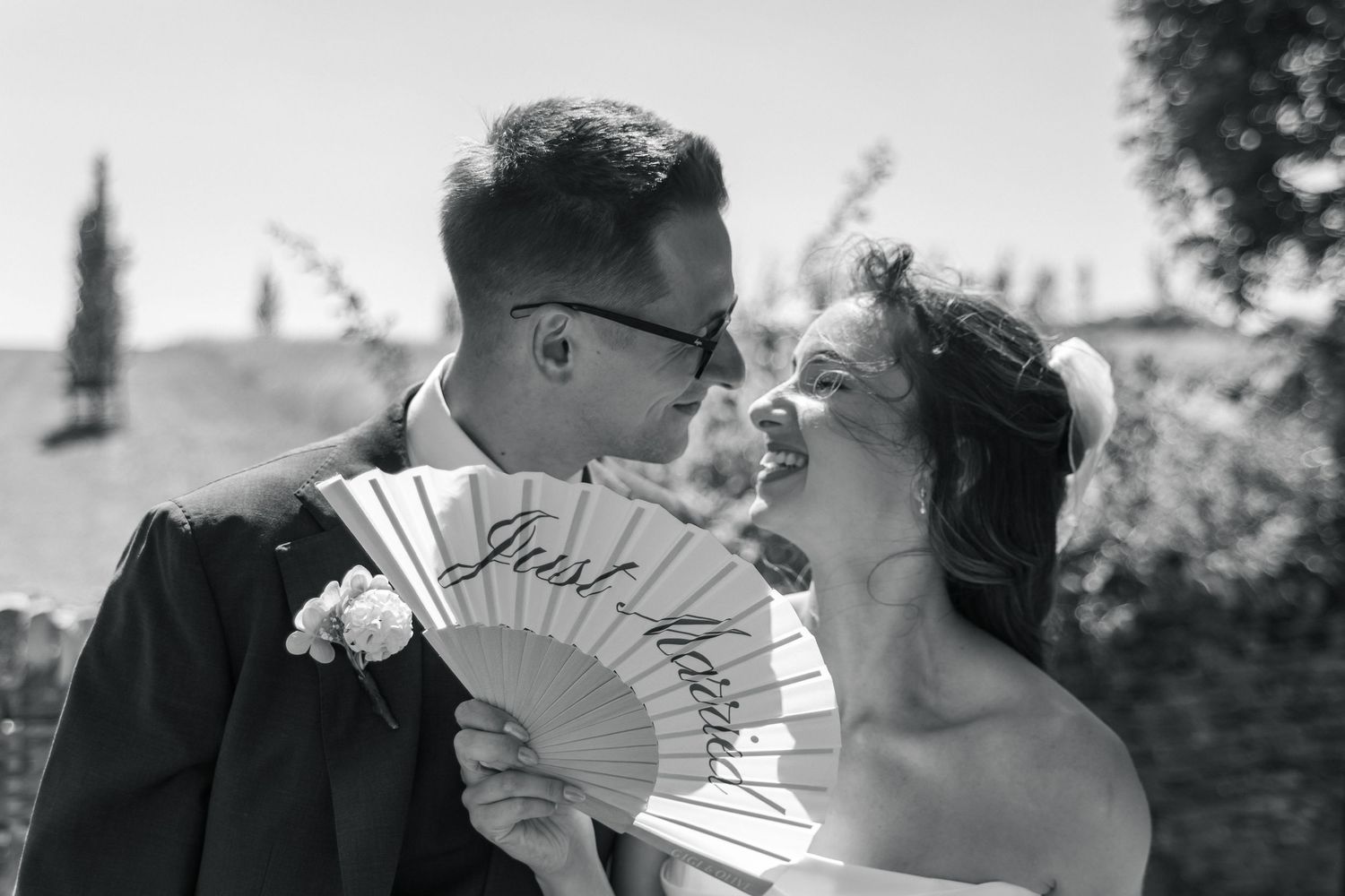 Black and white photo of newlyweds kissing behind a decorative fan reading Just Married on their wedding day.