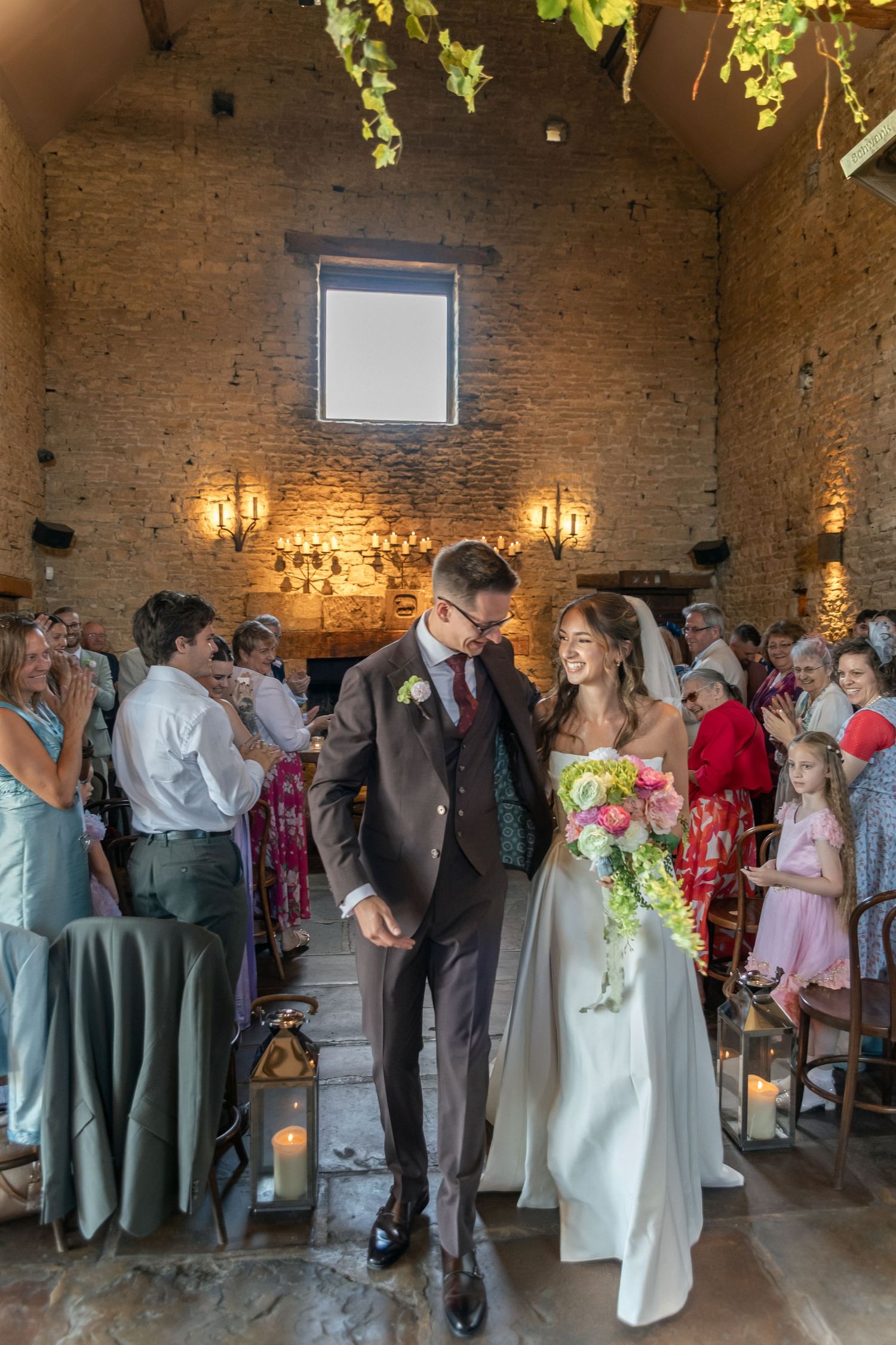 Bride and groom walk down the aisle in a rustic stone venue, guests applauding, bride holding a colorful bouquet.