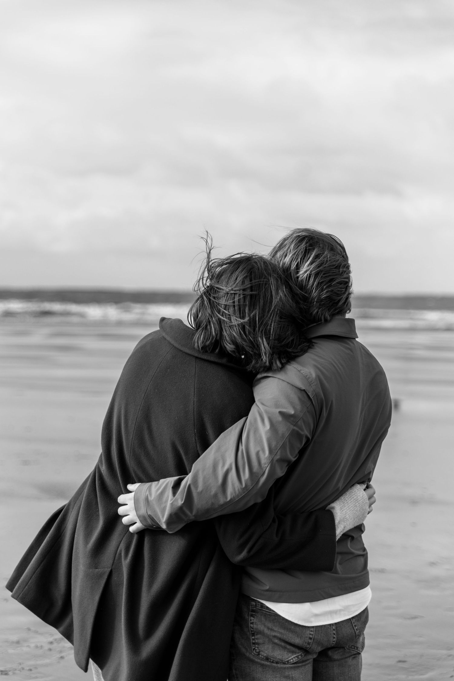 Two people embrace while looking out at the ocean waves on an overcast day.