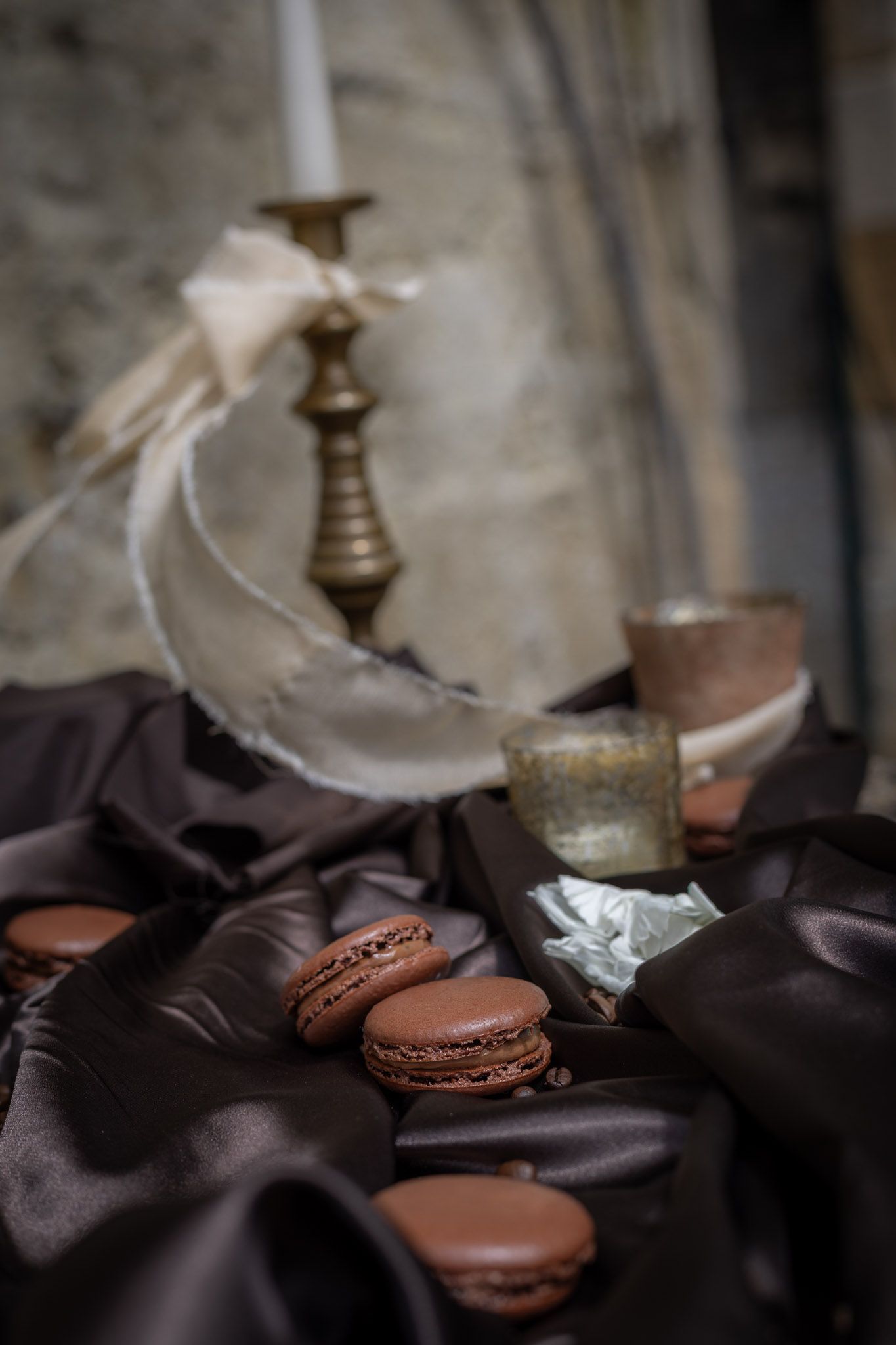 Chocolate macarons on dark satin fabric with a brass candlestick and vintage glass in moody still life.