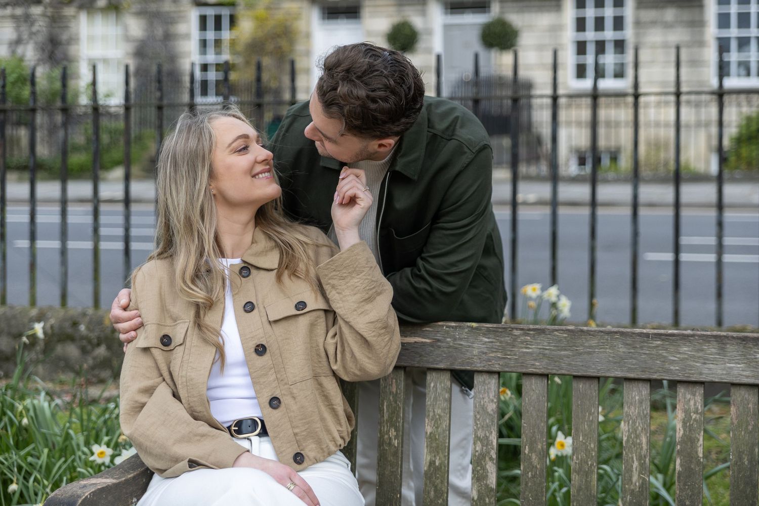 A smiling couple sitting on a bench outdoors, man kissing womans head in a romantic garden setting.