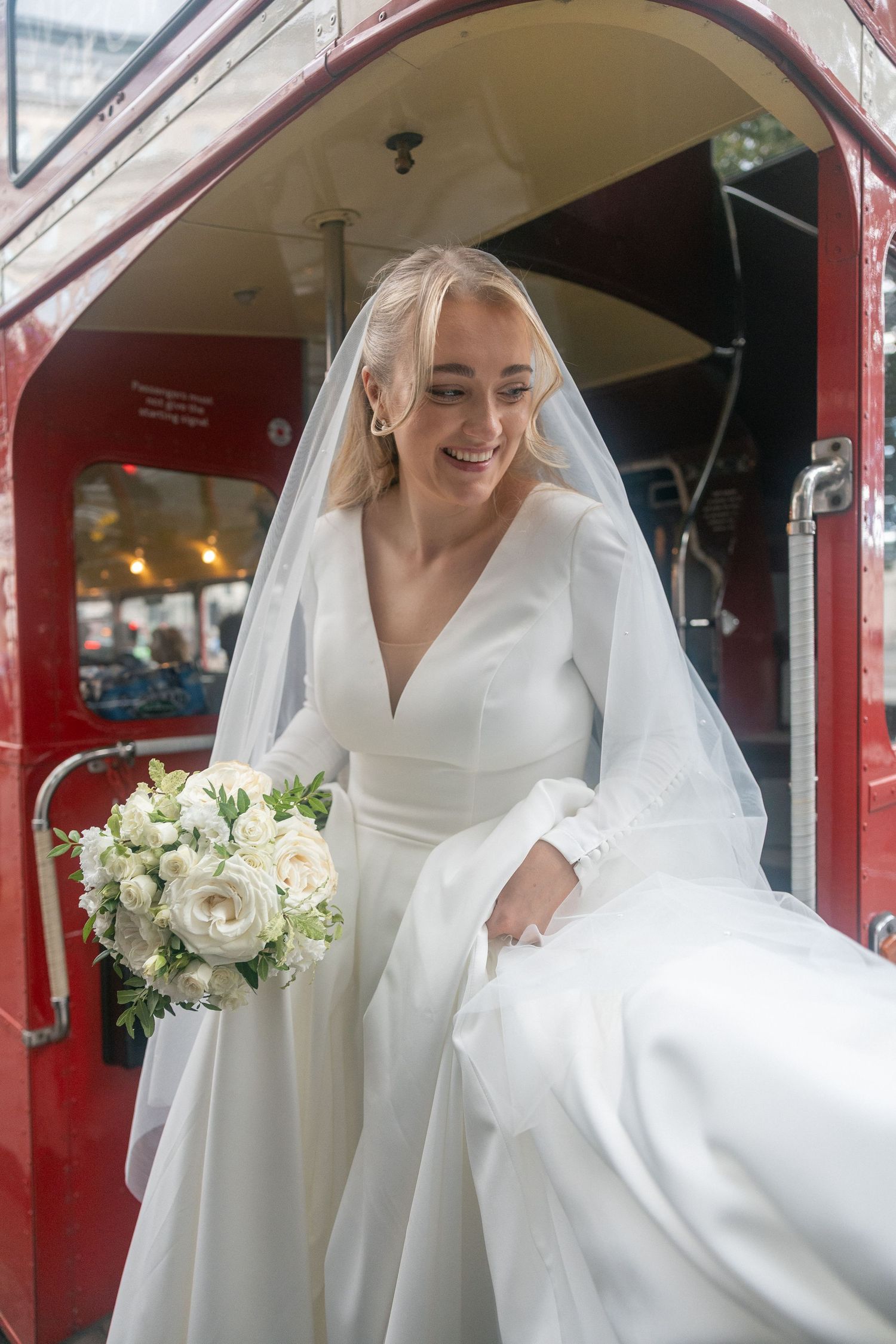 Smiling bride in white gown and veil holding bouquet of white roses stepping off a red double-decker bus.