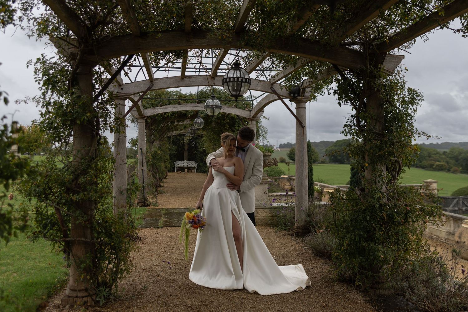 A bride in a flowing white gown stands beneath a rustic wooden arbor covered in greenery and flowers.
