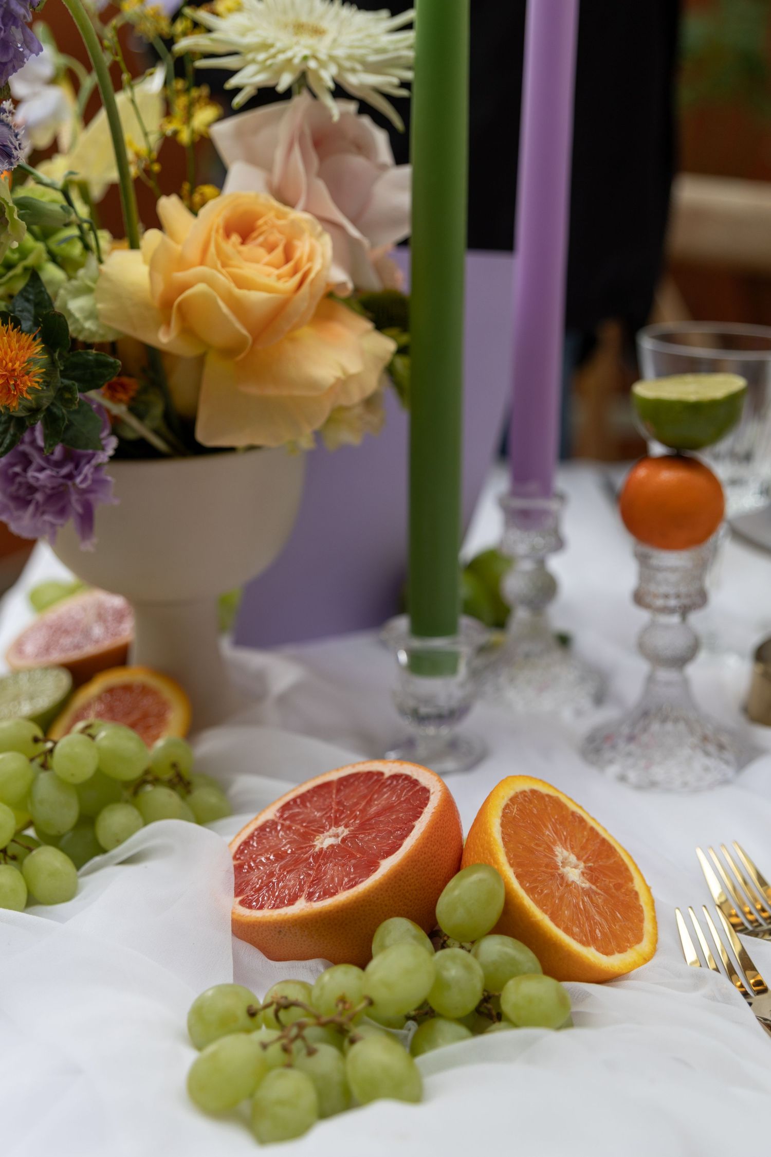 Elegant table setting with purple candles, fresh citrus fruits, green grapes and peach roses on white tablecloth.